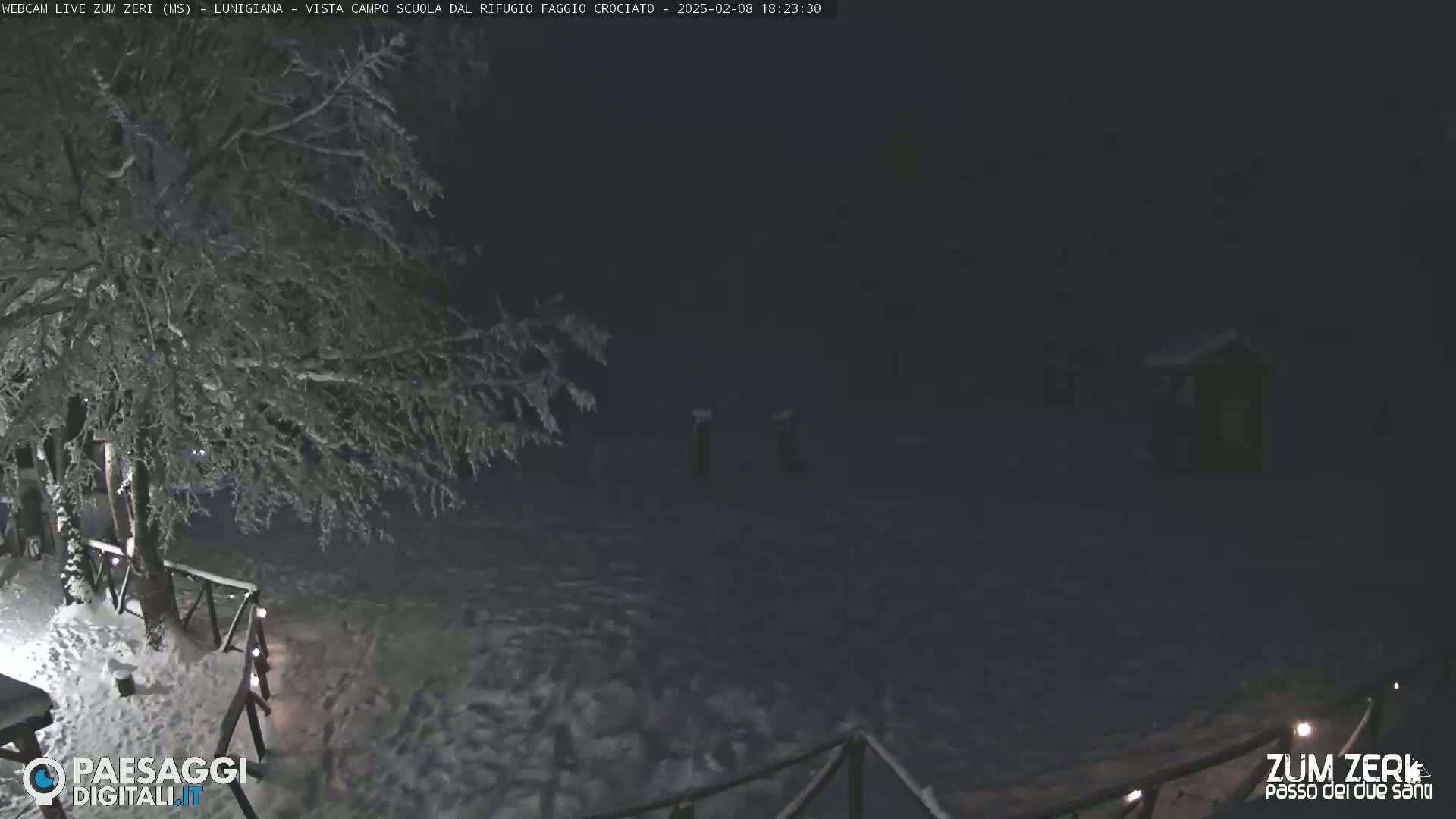 A snow-covered landscape at night, featuring a tree laden with snow, a wooden fence with small lights, and a dark structure in the distance.