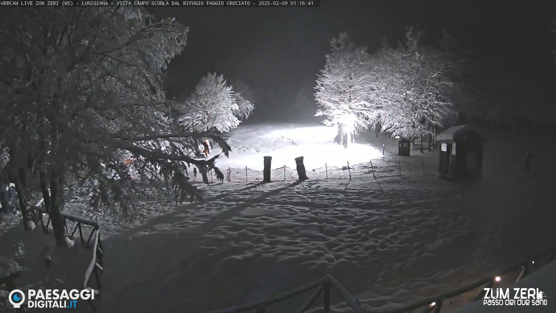 A snow-covered landscape at night, illuminated by artificial lights, shows snow-laden trees and small structures.