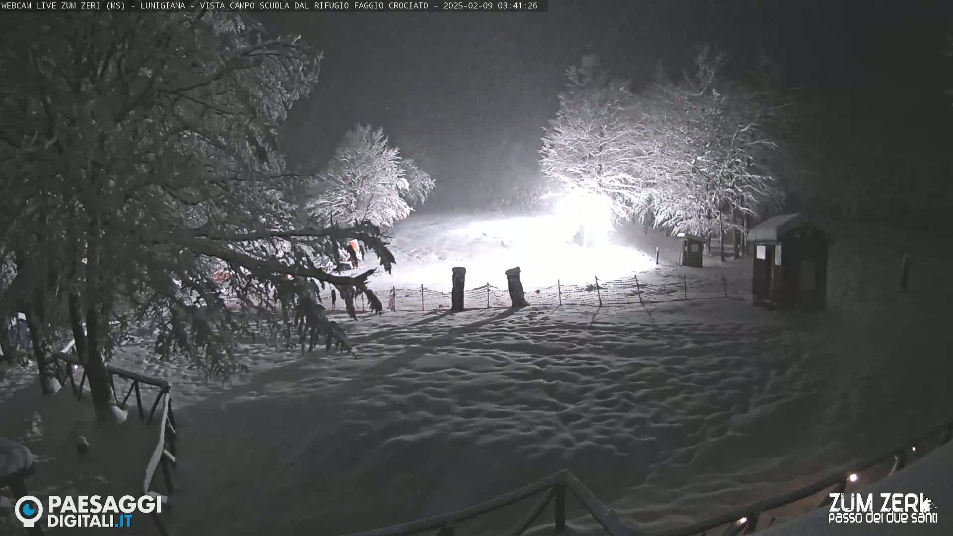 A snow-covered landscape at night, illuminated by artificial light, shows snow-laden trees and small structures.