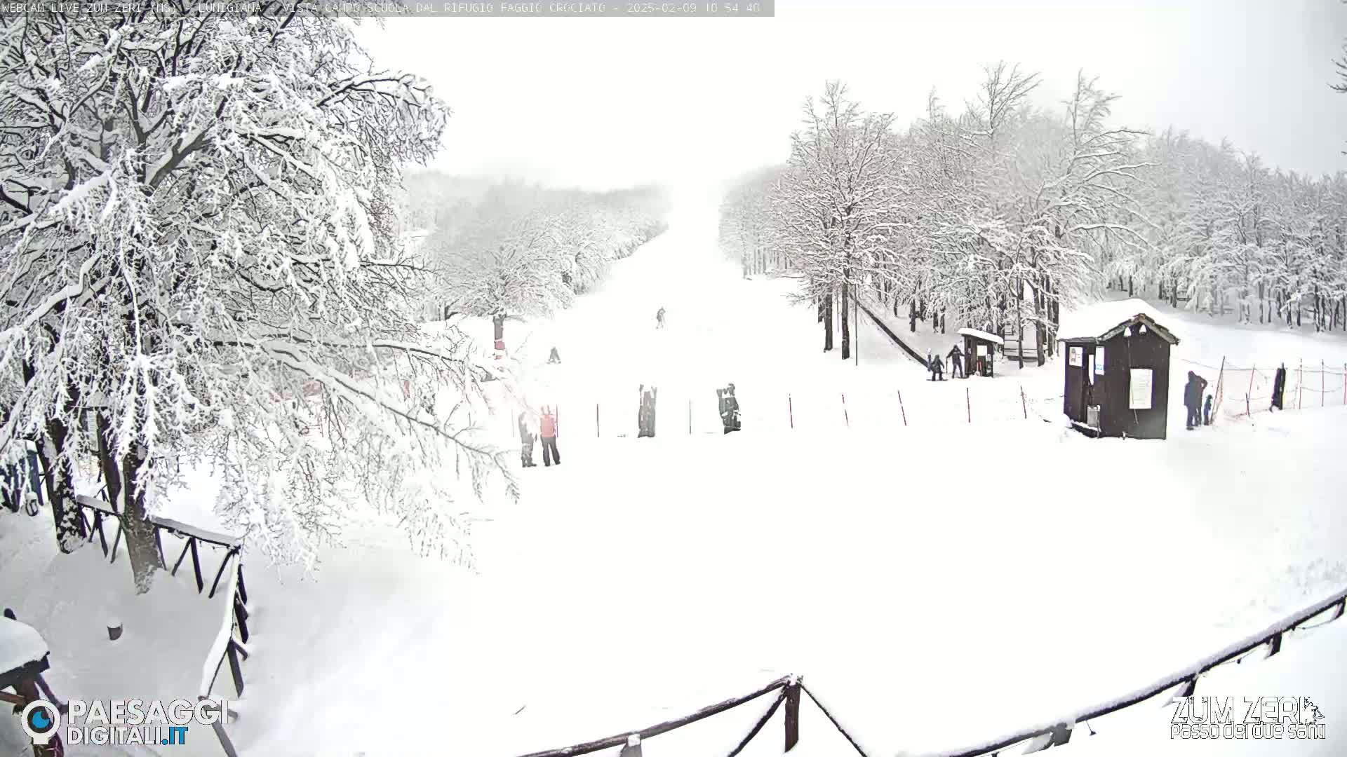 A snow-covered ski slope with several people and small structures is seen on a heavily overcast day.