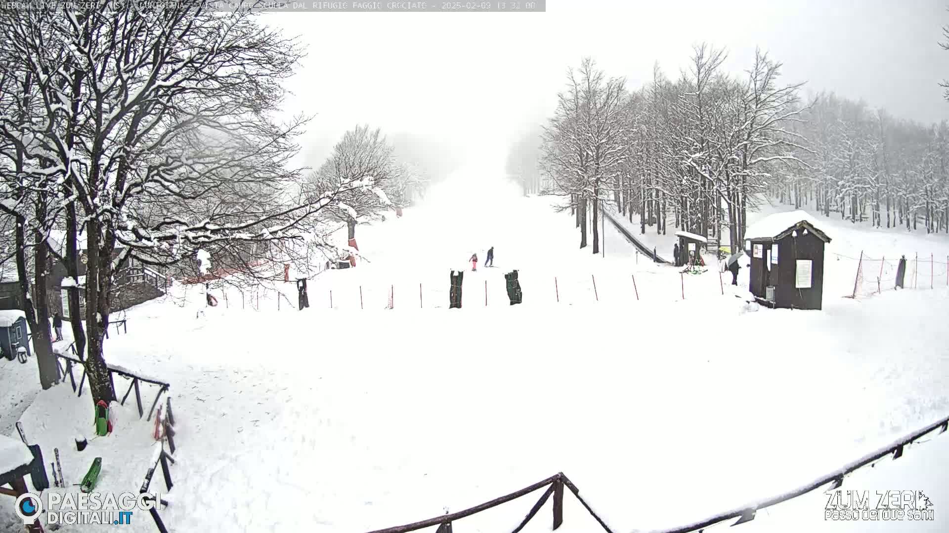 A snow-covered ski slope with several people skiing or snowboarding, surrounded by snow-laden trees in overcast weather.