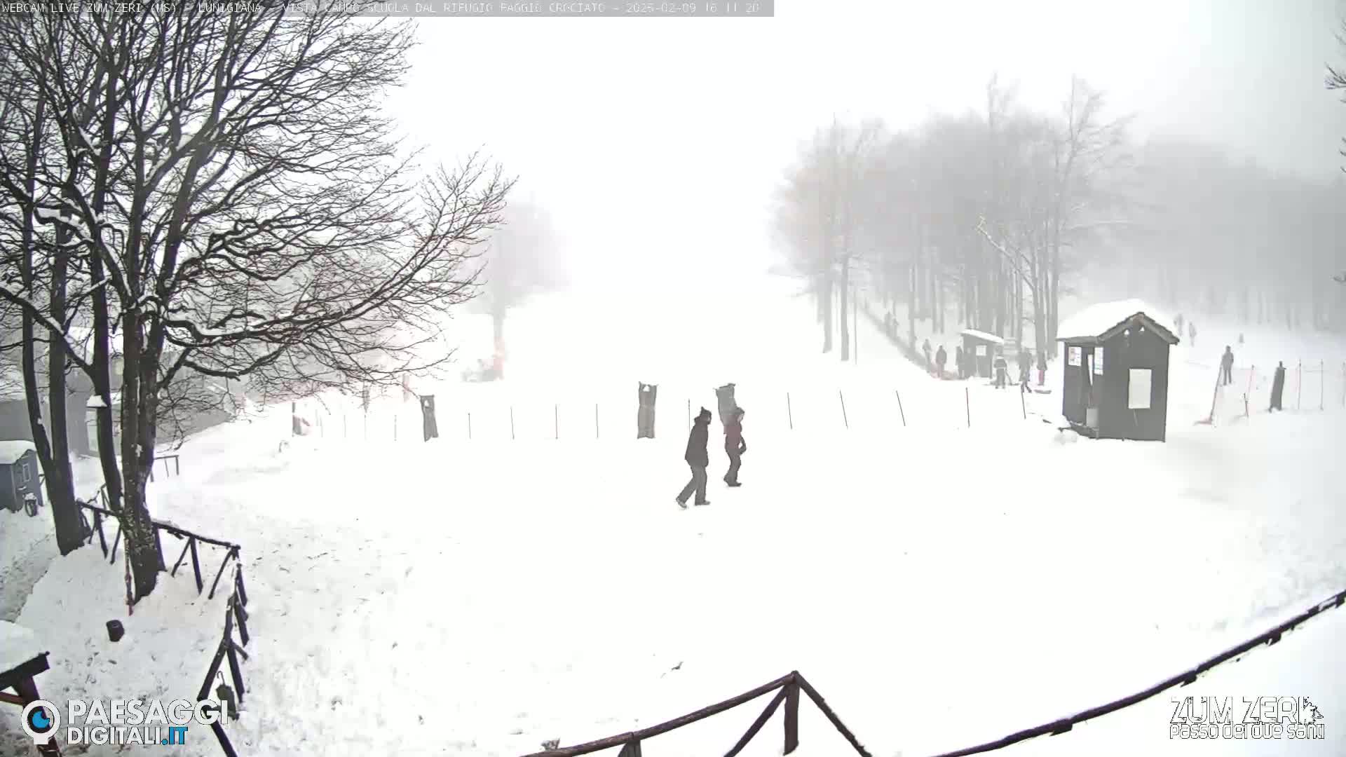 A snowy, foggy scene shows two people walking across a snow-covered area with bare trees and small buildings in the background.