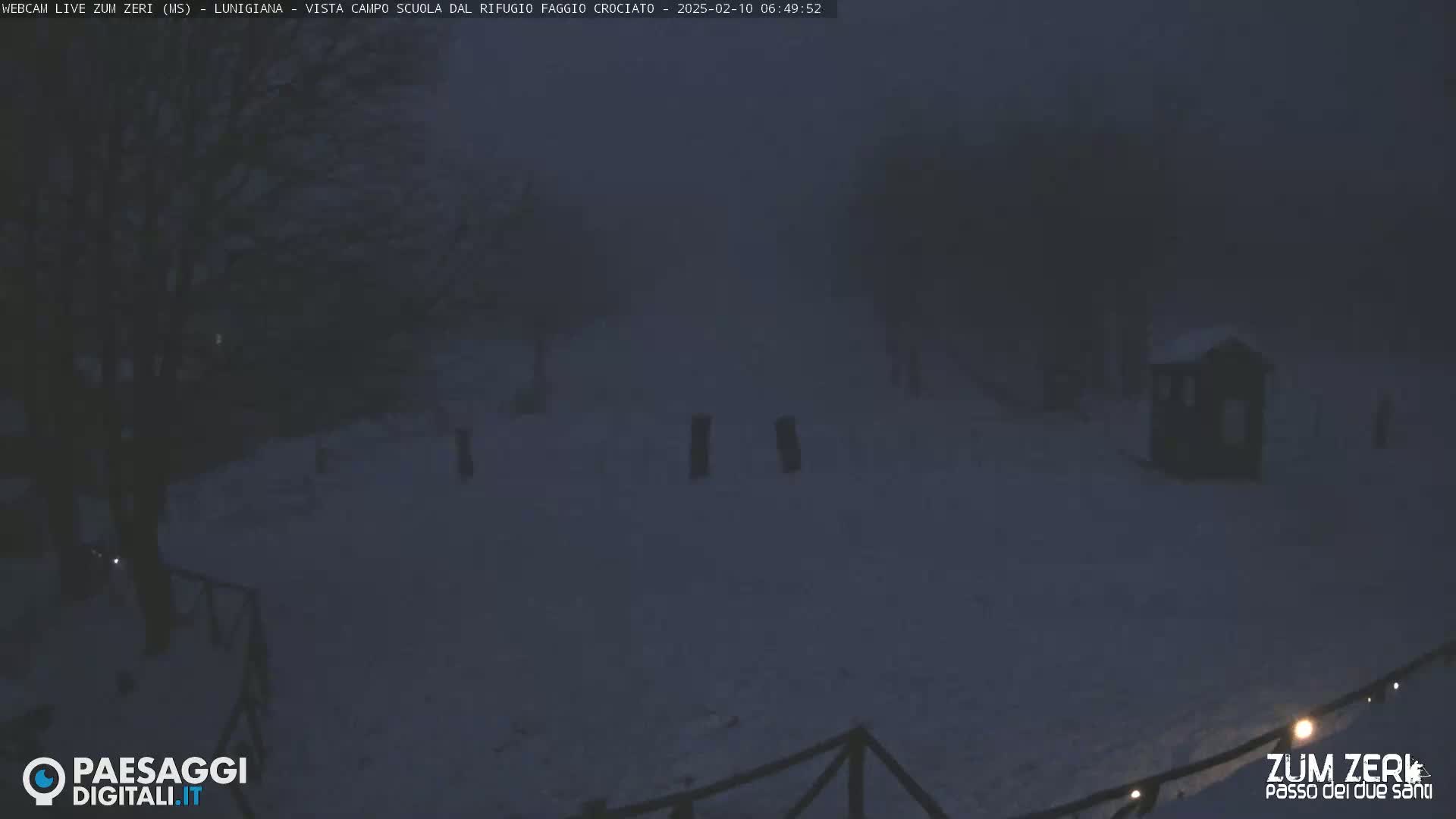 A snow-covered landscape at night is dimly lit, showing a small structure and trees under foggy conditions.
