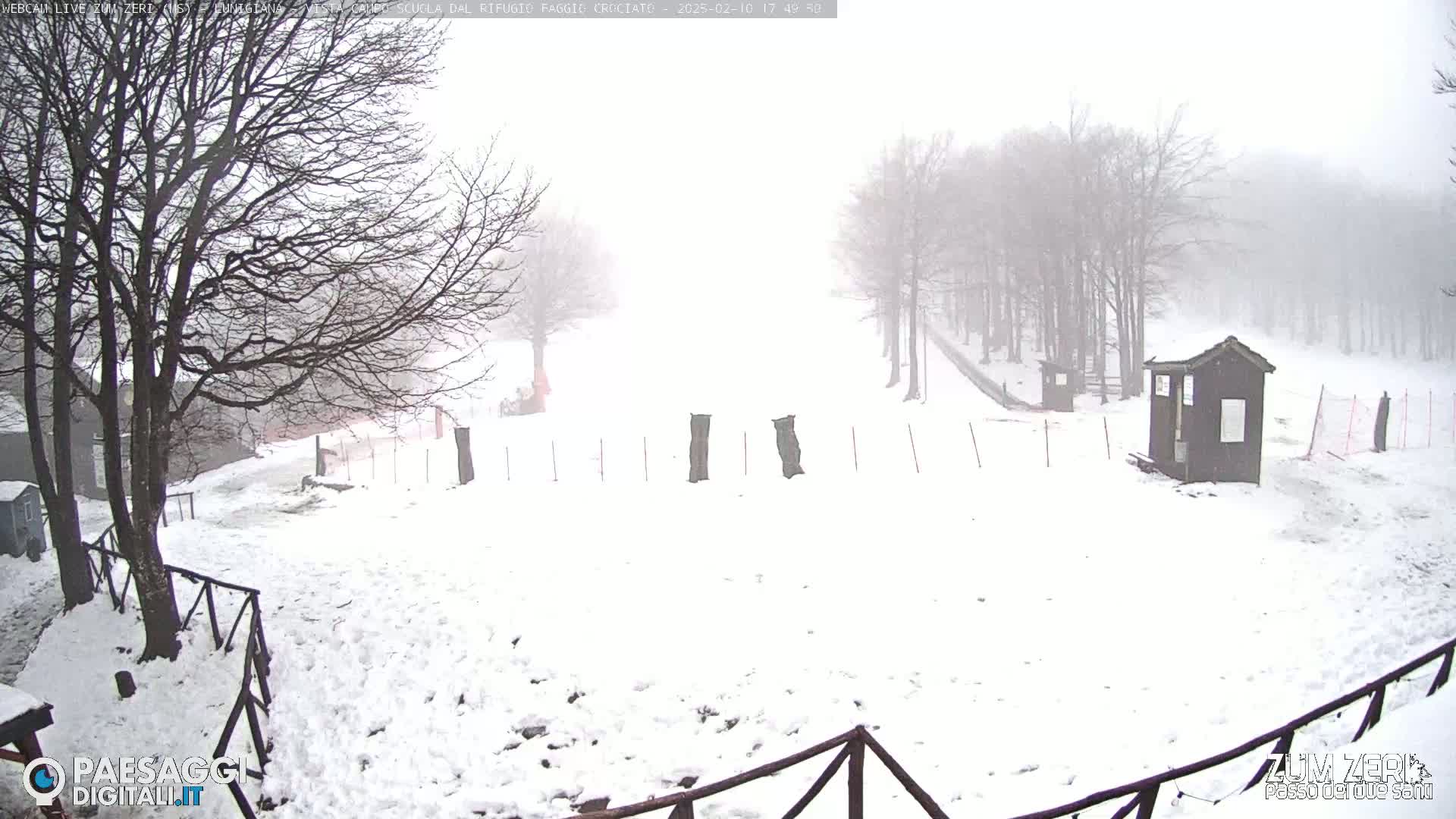 A snow-covered landscape with leafless trees in a foggy atmosphere features a small building and a fence.