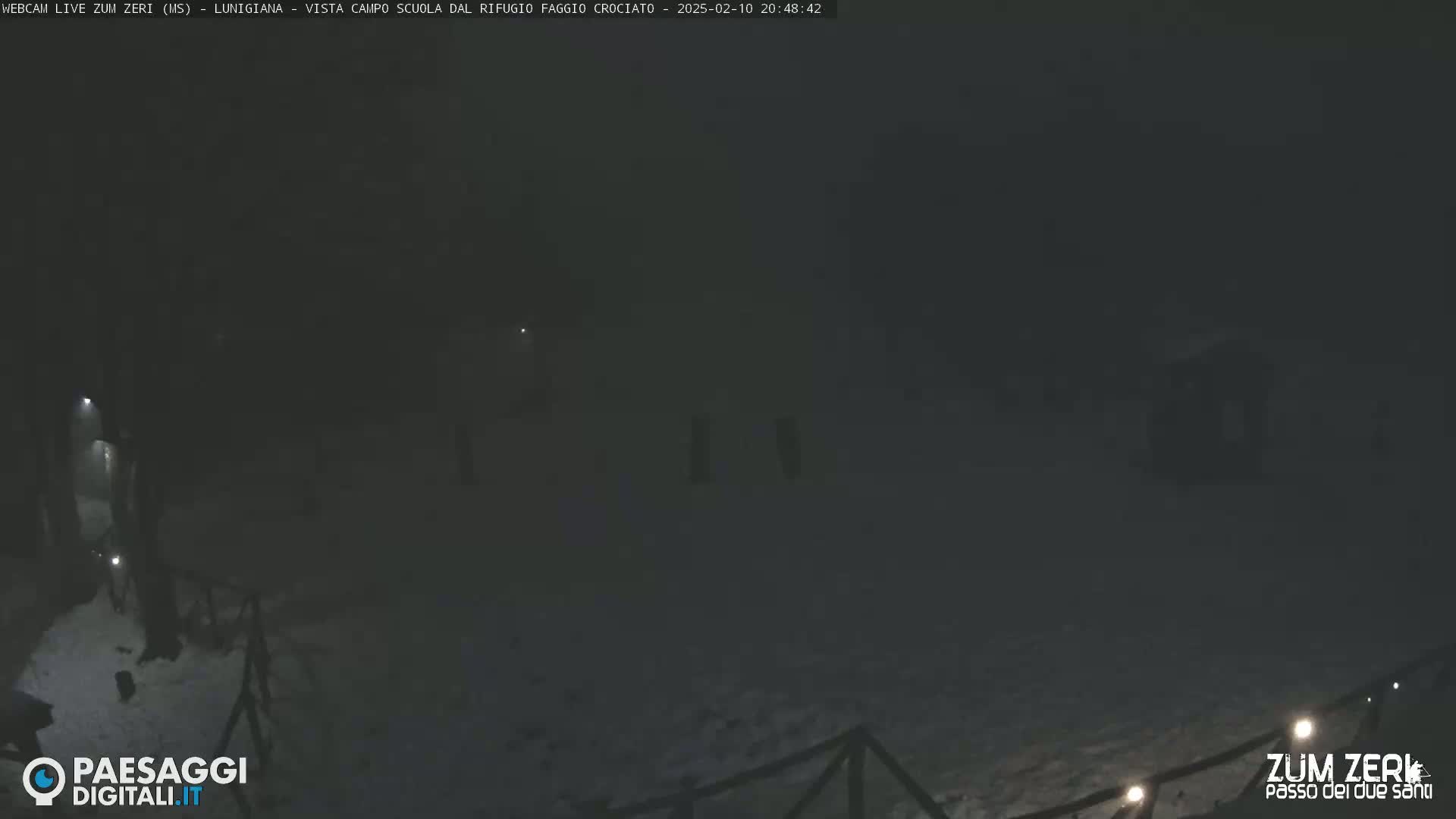 A nighttime, snow-covered outdoor scene is dimly lit, showing a dark area with a few indistinct structures and a fence.