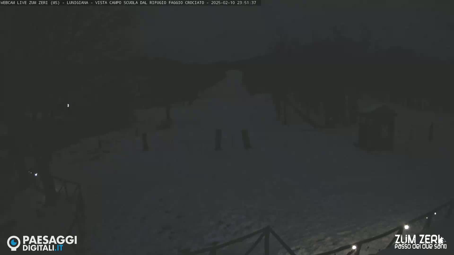A nighttime, snow-covered outdoor scene shows a sparsely lit area with several indistinct structures and a low fence.