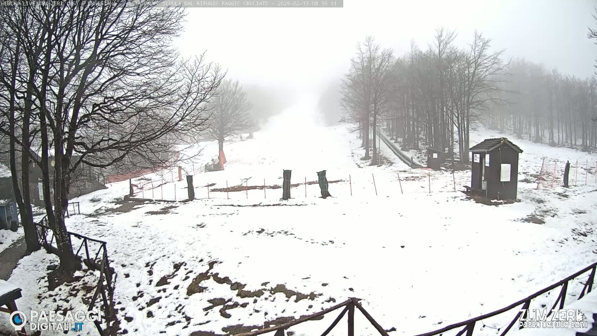 A snow-covered landscape with leafless trees in foggy conditions, showing a small building and a section of what appears to be a ski slope.