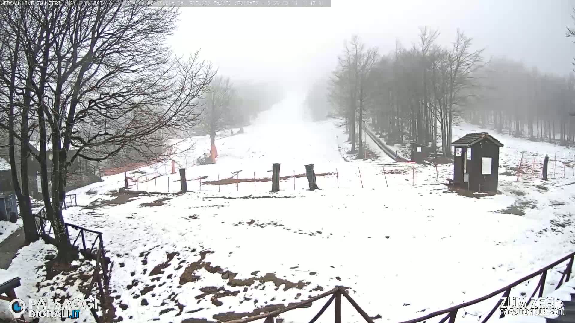 A snow-covered landscape with bare trees in foggy conditions shows a slight incline leading up to a misty background, with a small building and fencing visible in the foreground.
