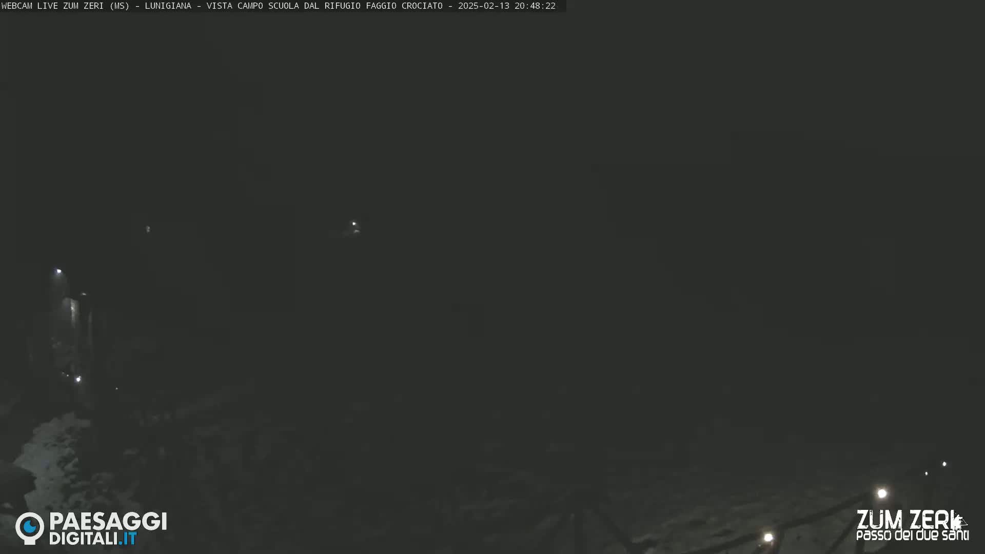 A dark, nighttime outdoor scene shows dimly lit areas of what appears to be snow-covered ground with a few small lights visible in the distance.