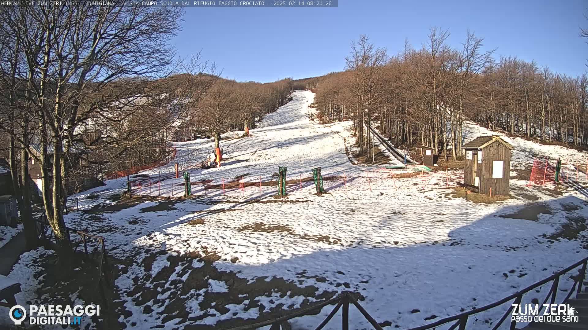 A snow-covered ski slope with bare trees and a small wooden building under a clear blue sky.
