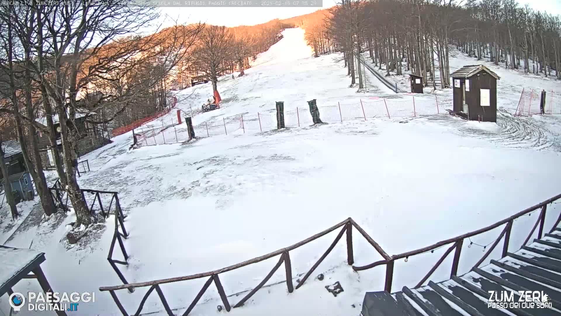 A snow-covered ski slope is seen from a high vantage point on a sunny day, with a wooden fence and small buildings in the foreground.