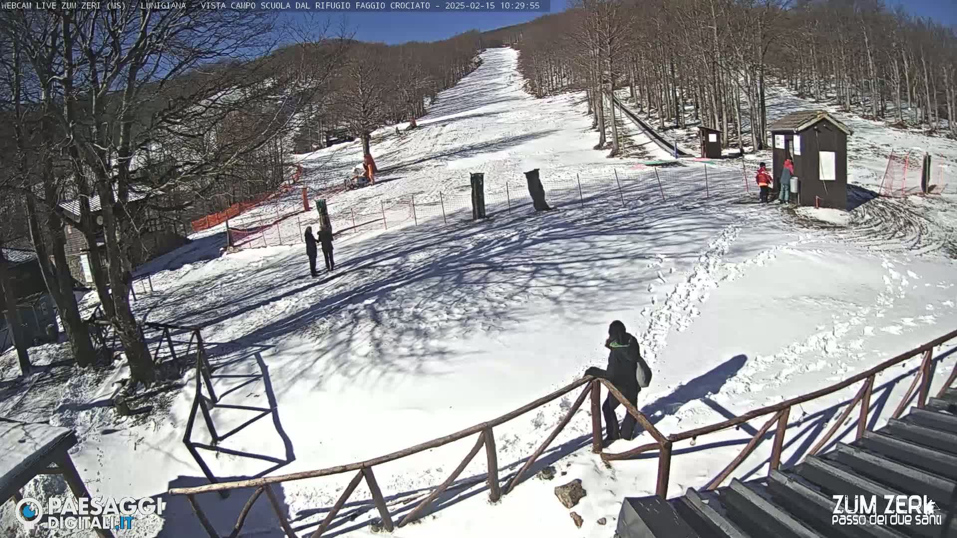 A snow-covered ski slope with several people near a small building under a sunny sky.