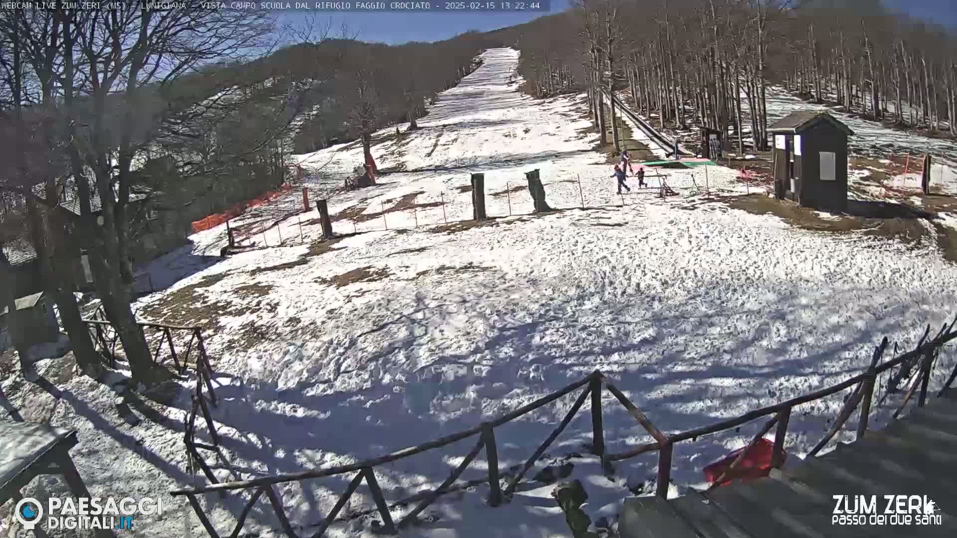 A snow-covered ski slope with several people near a small building, under a sunny sky.