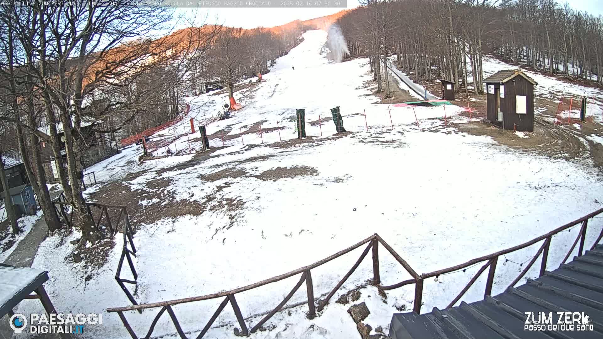 A snow-covered ski slope with a few small buildings and a wooden fence is visible under a partly sunny sky.