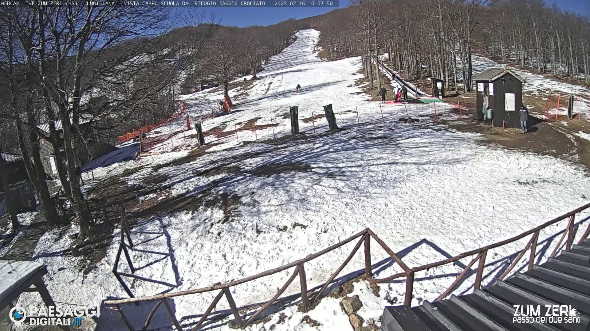 A snow-covered ski slope with several people near a small building on a sunny day.