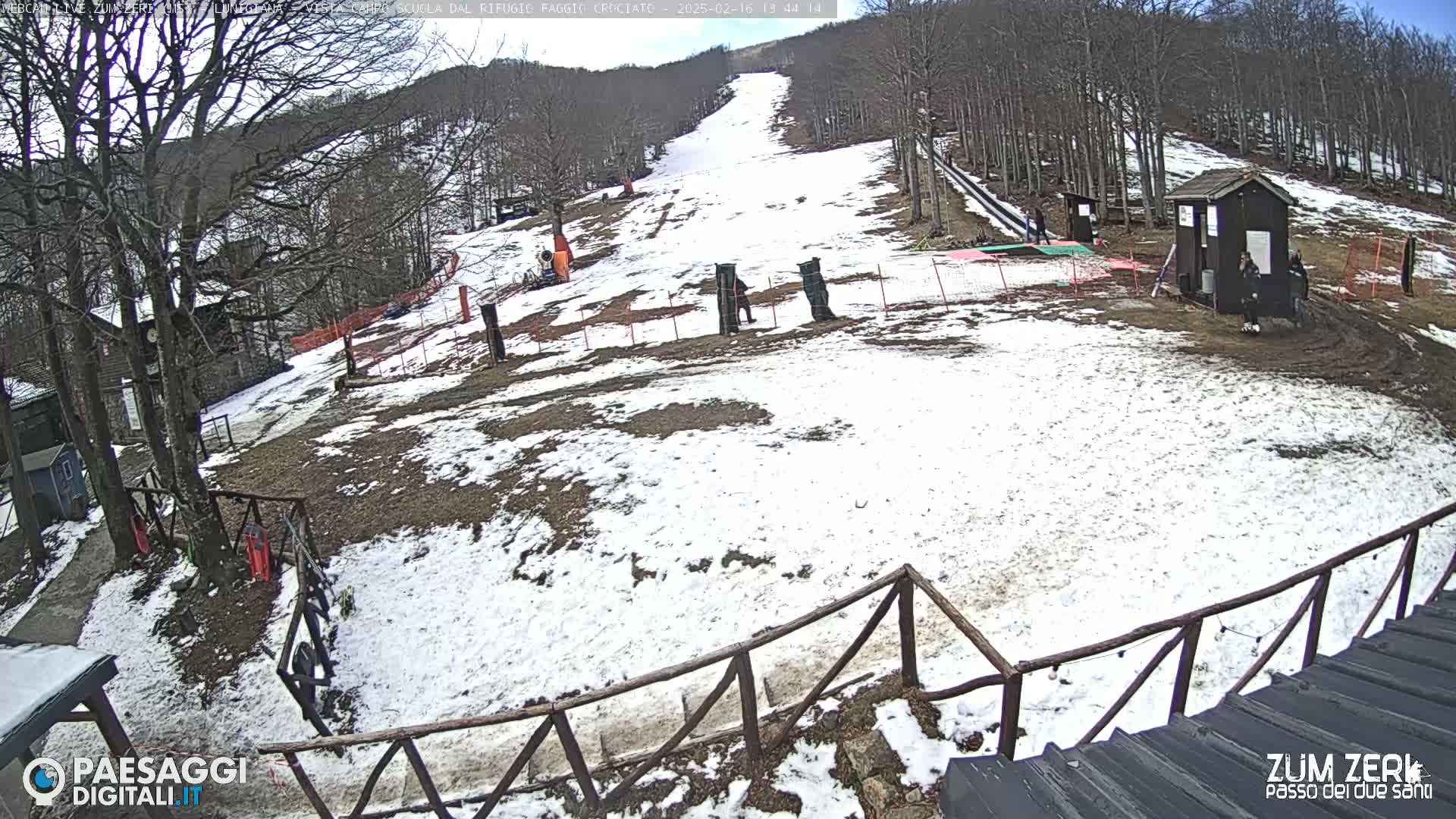 A partially snow-covered ski slope is visible, with a few people near a small building at the bottom, under a mostly clear sky.