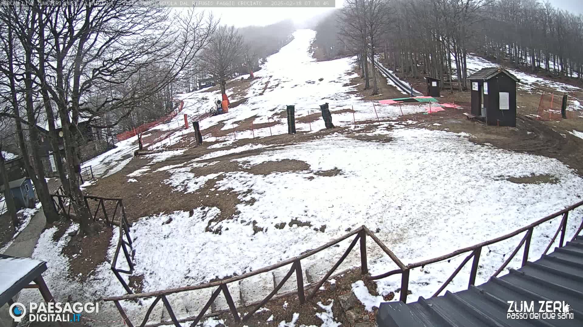A snowy, overcast scene shows a gently sloping hill with a wooden fence and small structures, surrounded by bare trees.