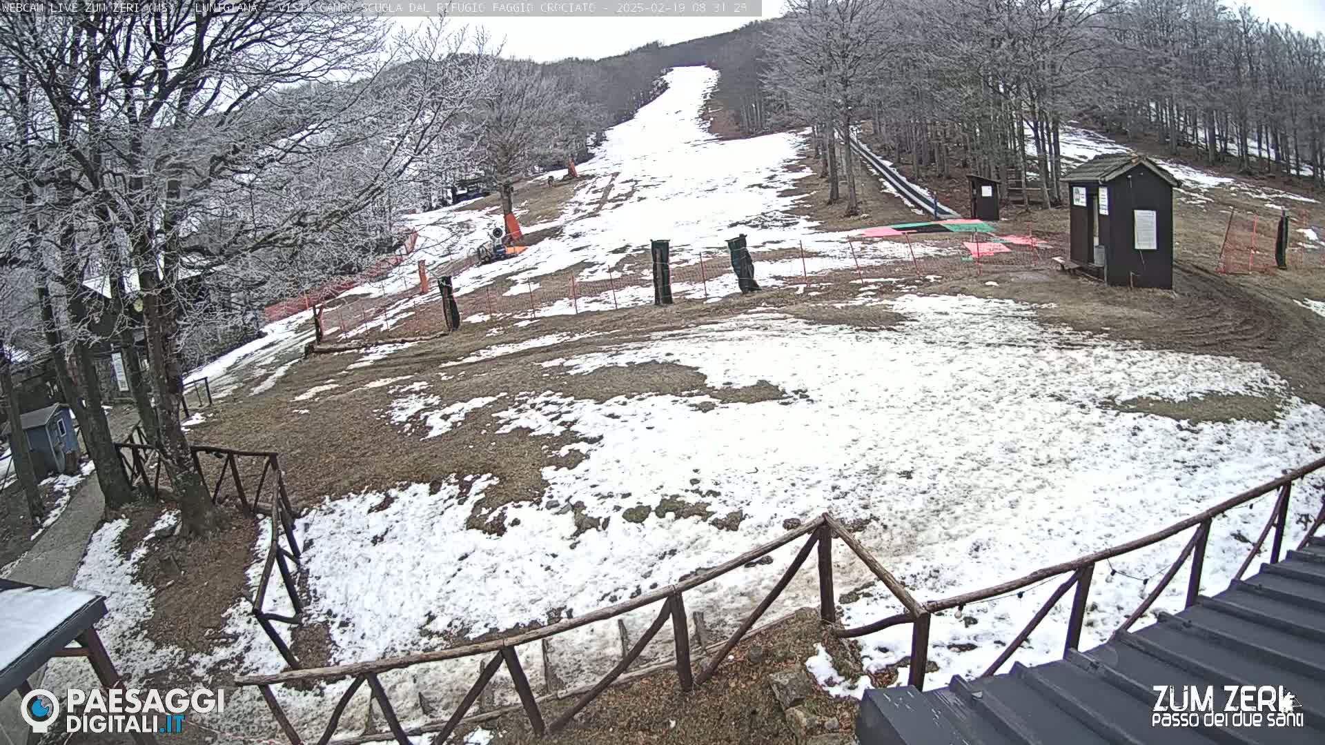 A snow-dusted ski slope is visible, bordered by trees and wooden fencing, with small structures nearby under an overcast sky.