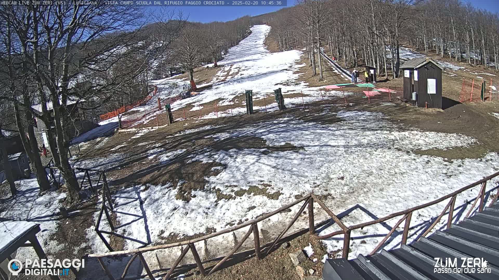 A snow-covered ski slope is seen on a sunny day, with a small building and several people nearby, all surrounded by bare trees.