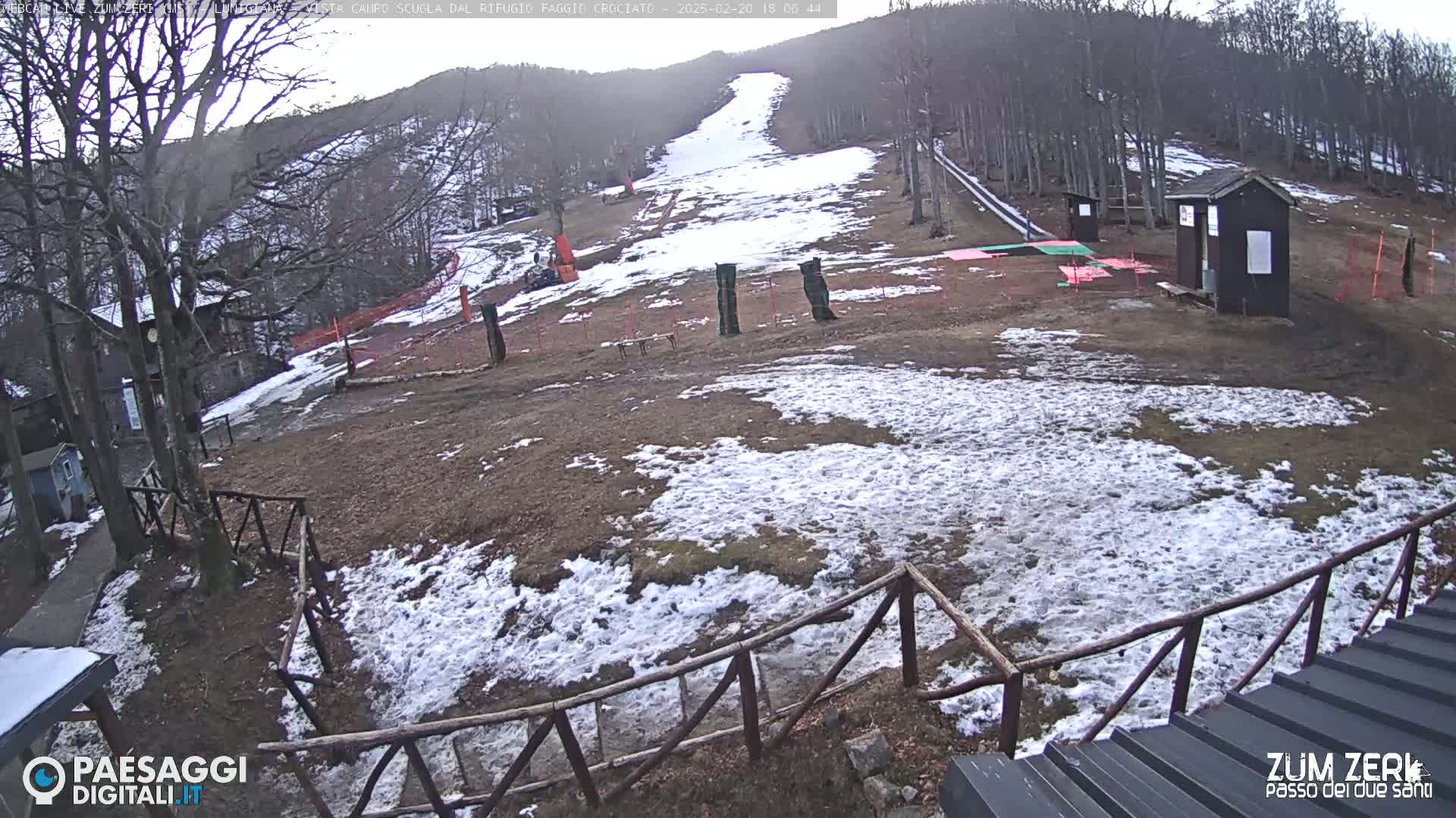 A partially snow-covered ski slope is viewed from behind a wooden fence, with several small buildings and bare trees visible in the background on an overcast day.