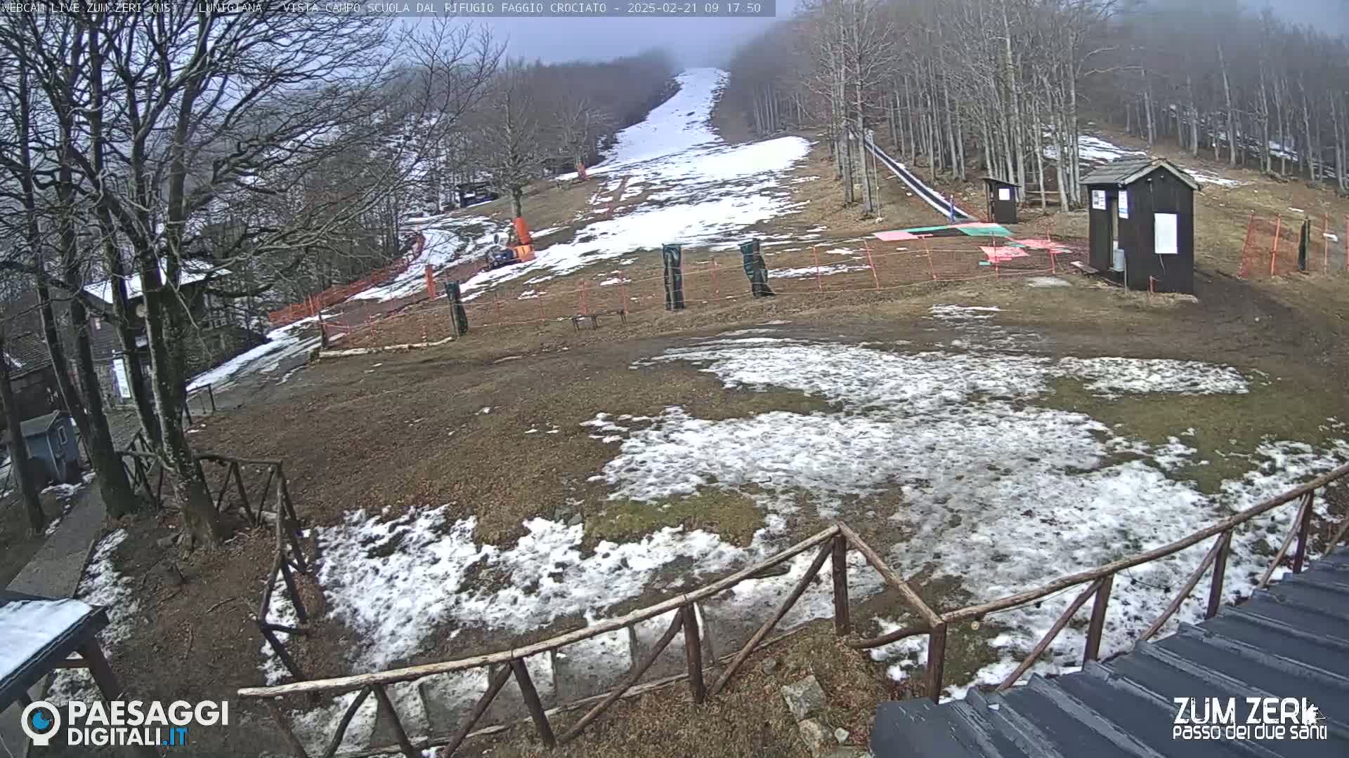 A snow-dusted ski slope with several small buildings and a wooden fence is visible under an overcast sky.