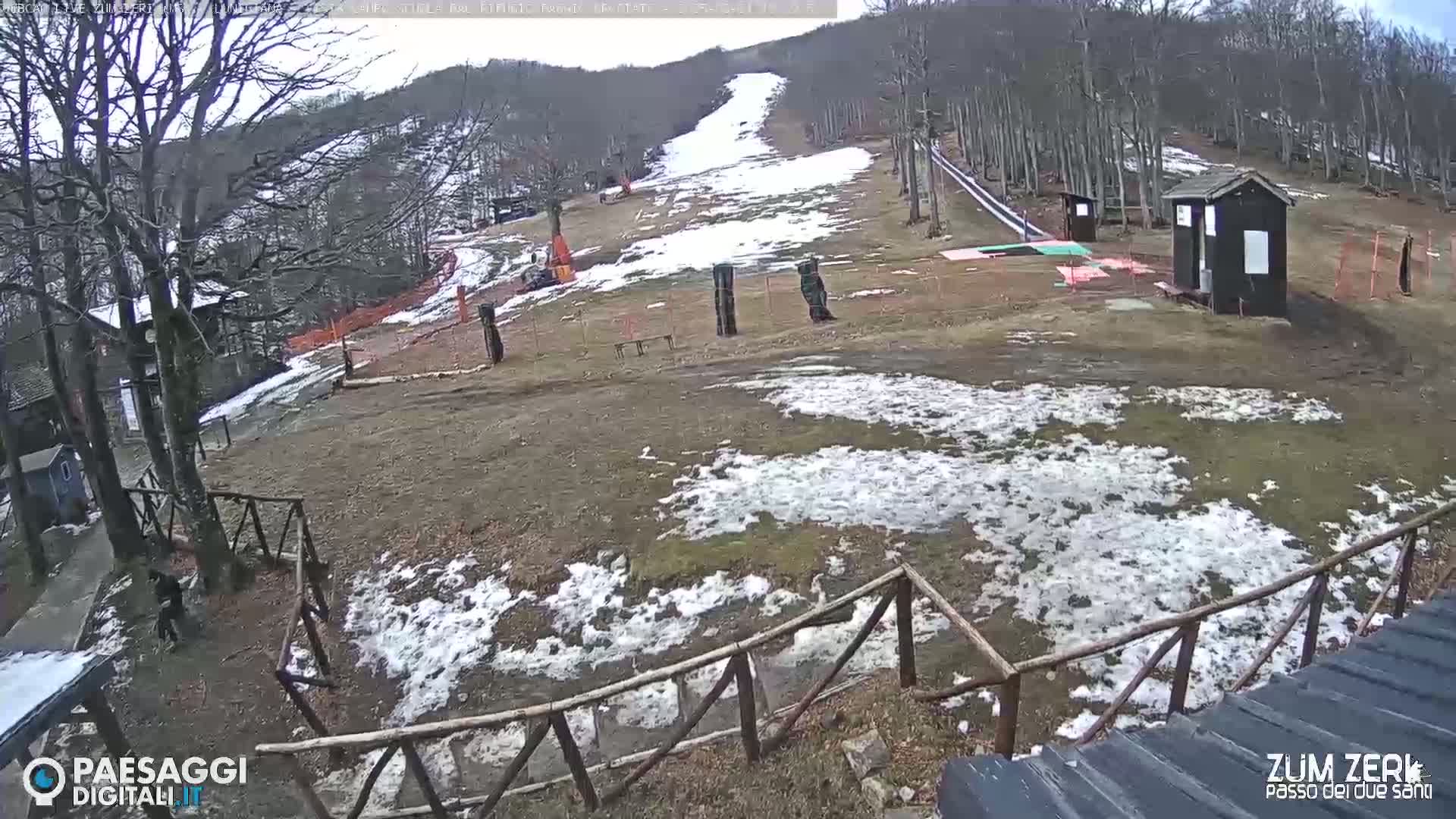 A mostly barren ski slope with patches of melting snow is visible, surrounded by trees under a cloudy sky.