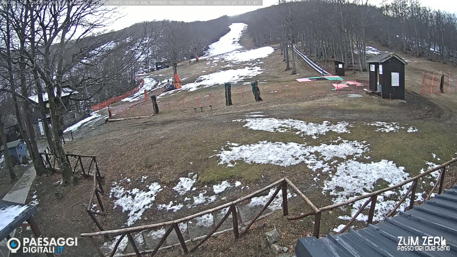 A mostly snow-free ski slope with patchy snow cover and a few small buildings, viewed from above on an overcast day.