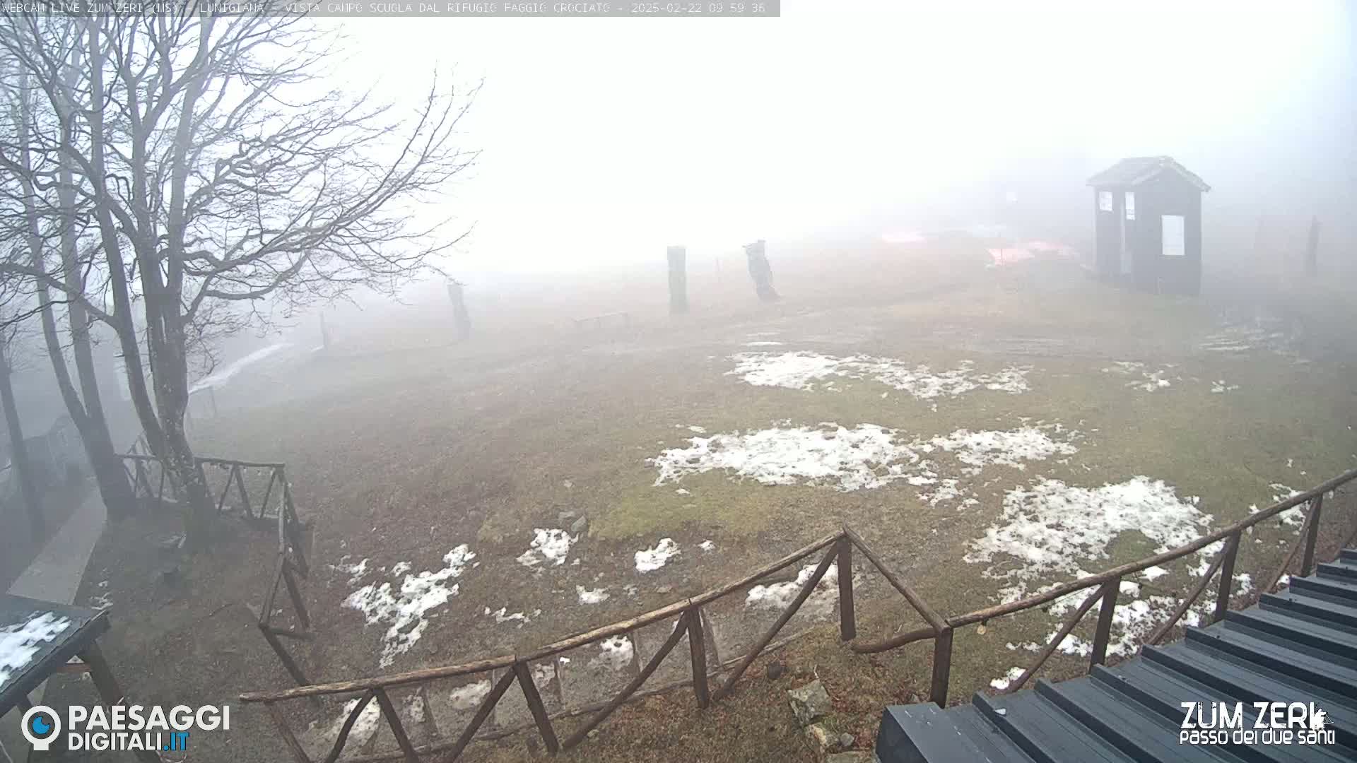 A foggy, snow-dusted field is enclosed by a wooden fence, with a small shed visible in the background and leafless trees in the foreground.