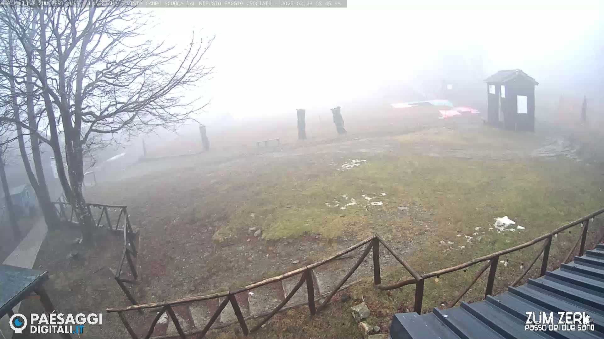 A foggy, outdoor scene shows a sparsely vegetated area with a wooden fence, a small building, and bare trees.