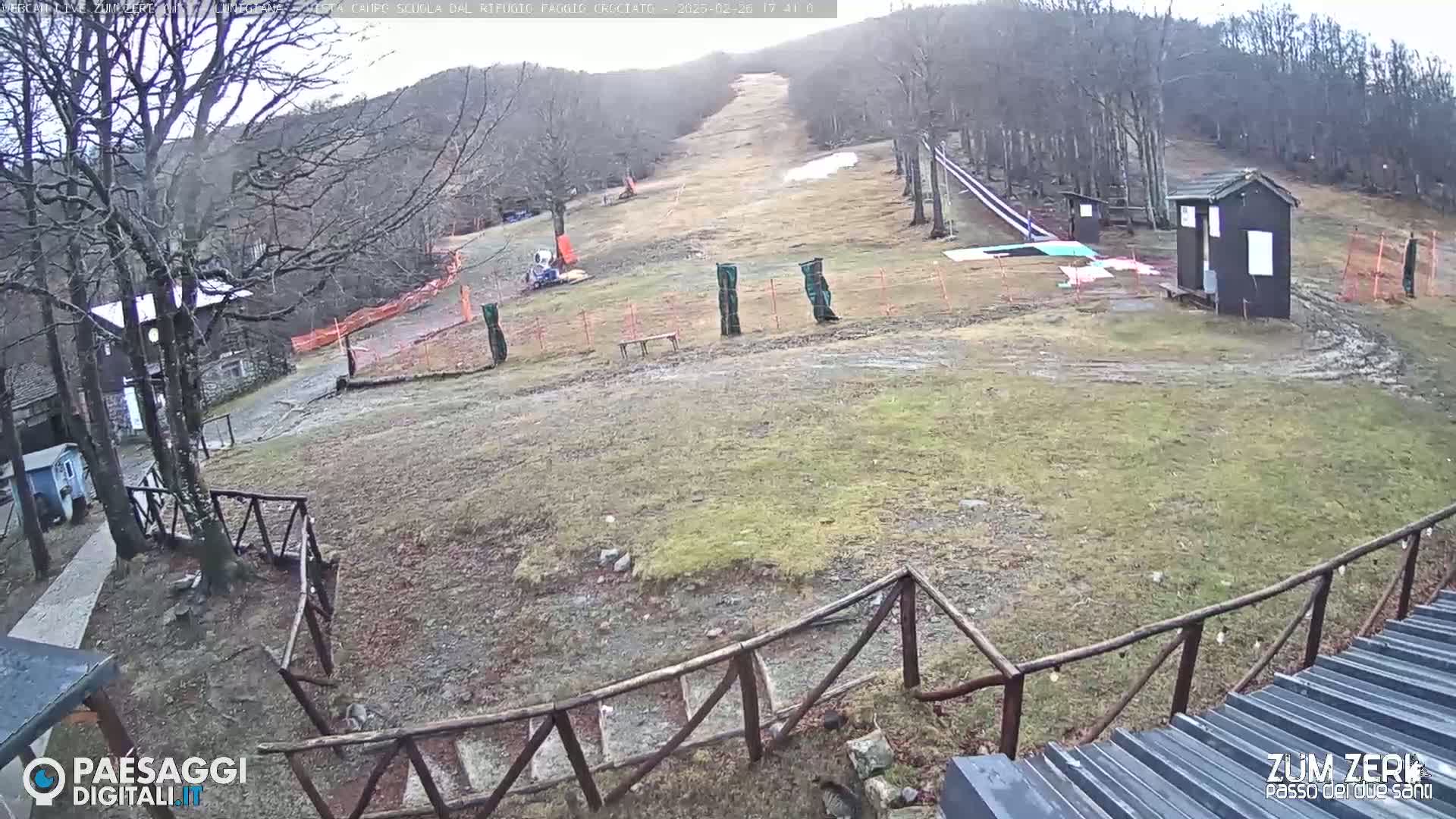 A mostly barren ski slope with a small building and wooden fencing is visible on an overcast day.