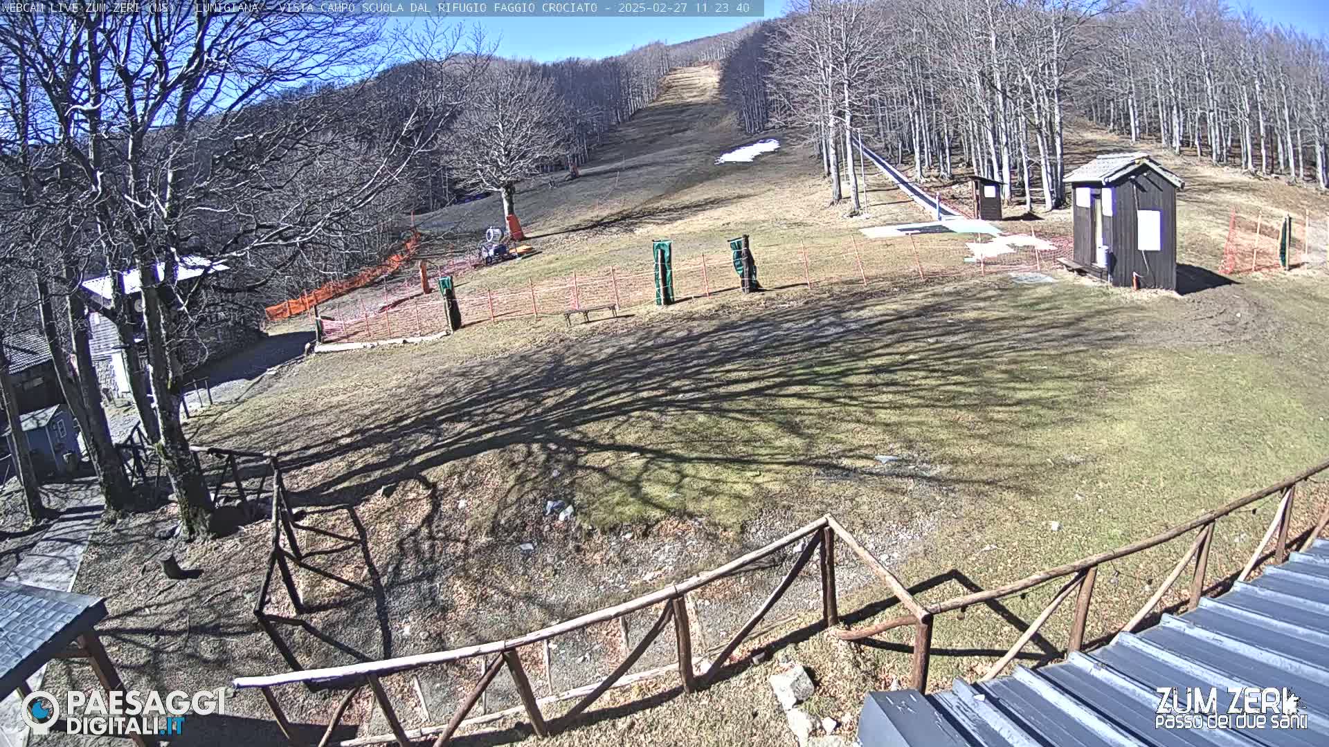 A sunny outdoor scene shows a grassy area with a ski slope, several small buildings, and bare trees under a clear blue sky.