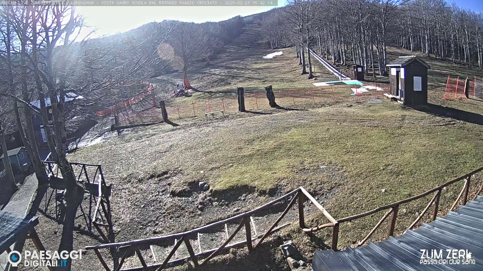 A sunny, snow-free ski slope is partially visible, with a small wooden building and several fences present; bare trees line the hillside in the background.