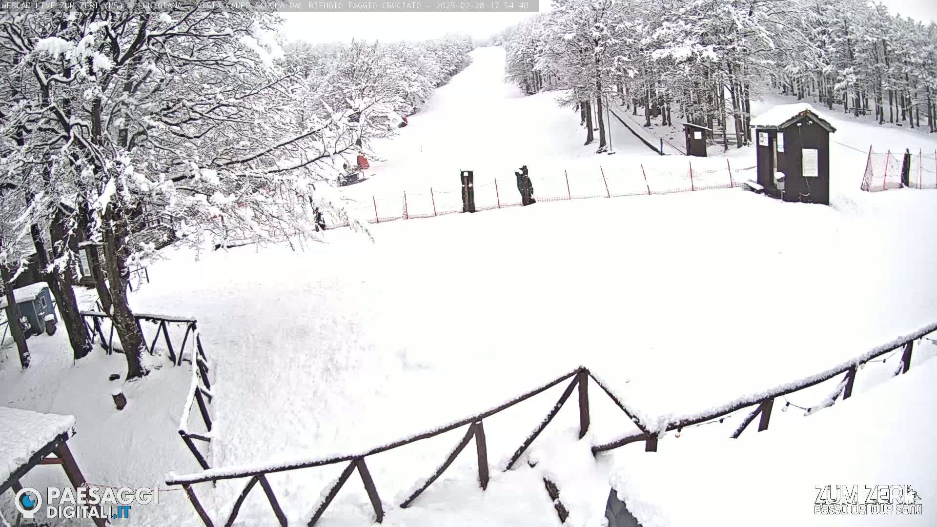 A snow-covered ski slope with a small building and wooden fence is visible, surrounded by snow-laden trees in a snowy landscape.