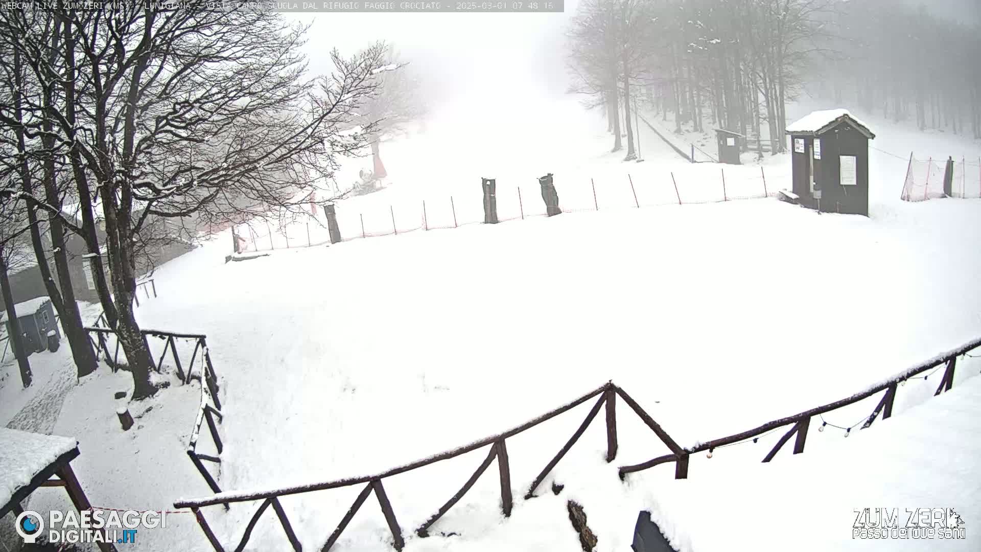 A snow-covered area with wooden fences, trees, and a small building in a foggy atmosphere.