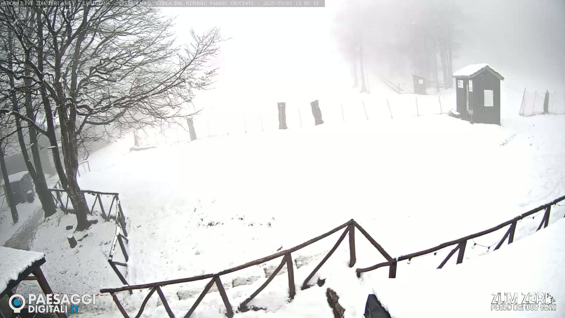 A snow-covered landscape is seen through a hazy fog, featuring a wooden fence, bare trees, and a small building.