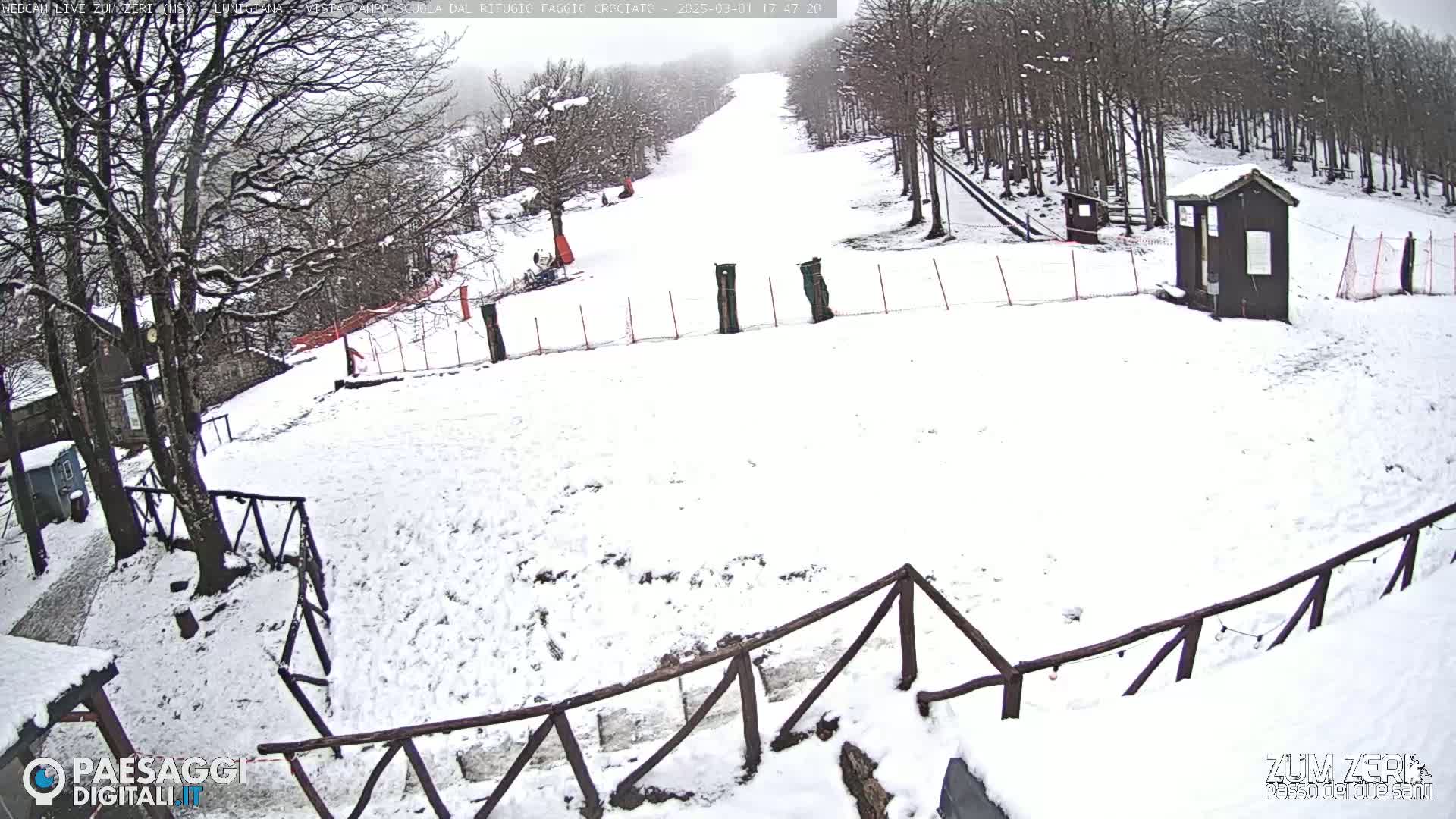 A snow-covered ski slope is visible, with a small building and wooden fences, under an overcast sky.