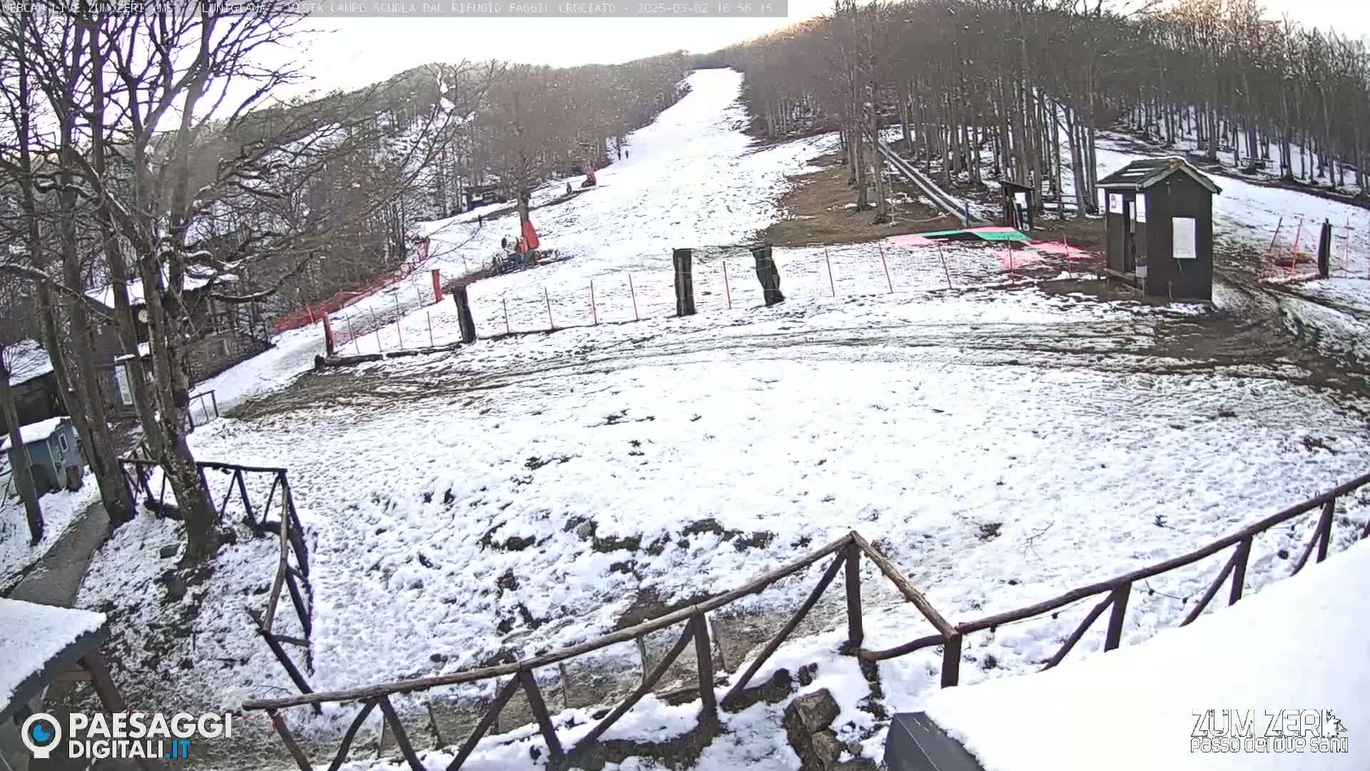 A snow-covered ski slope is visible, with a small building and wooden fences in the foreground, under a partly sunny sky.