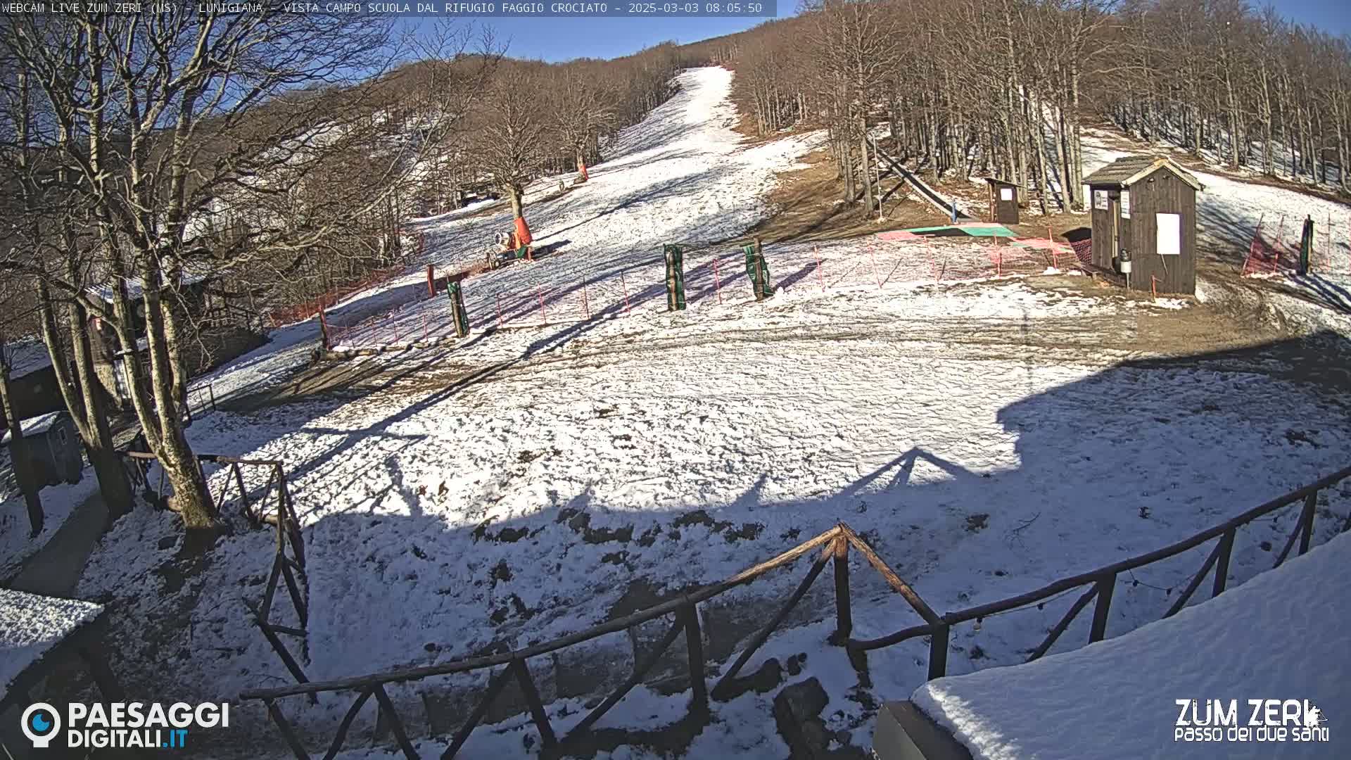 A snow-covered ski slope is seen on a sunny day, with a small building and some wooden fencing in the foreground.