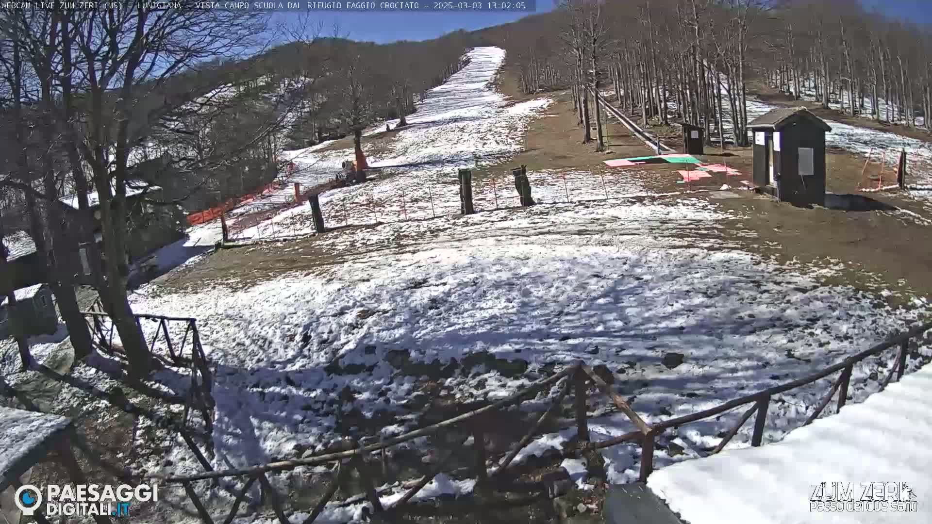 A snow-dusted ski slope is visible, with a small building and surrounding bare trees under a sunny sky.