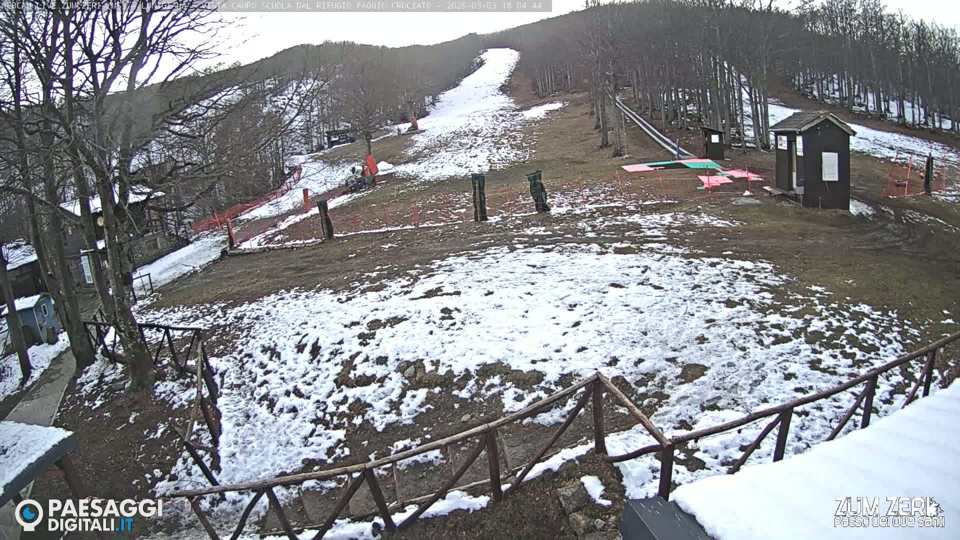 A partially snow-covered ski slope with a small building and wooden fence is visible on an overcast day.