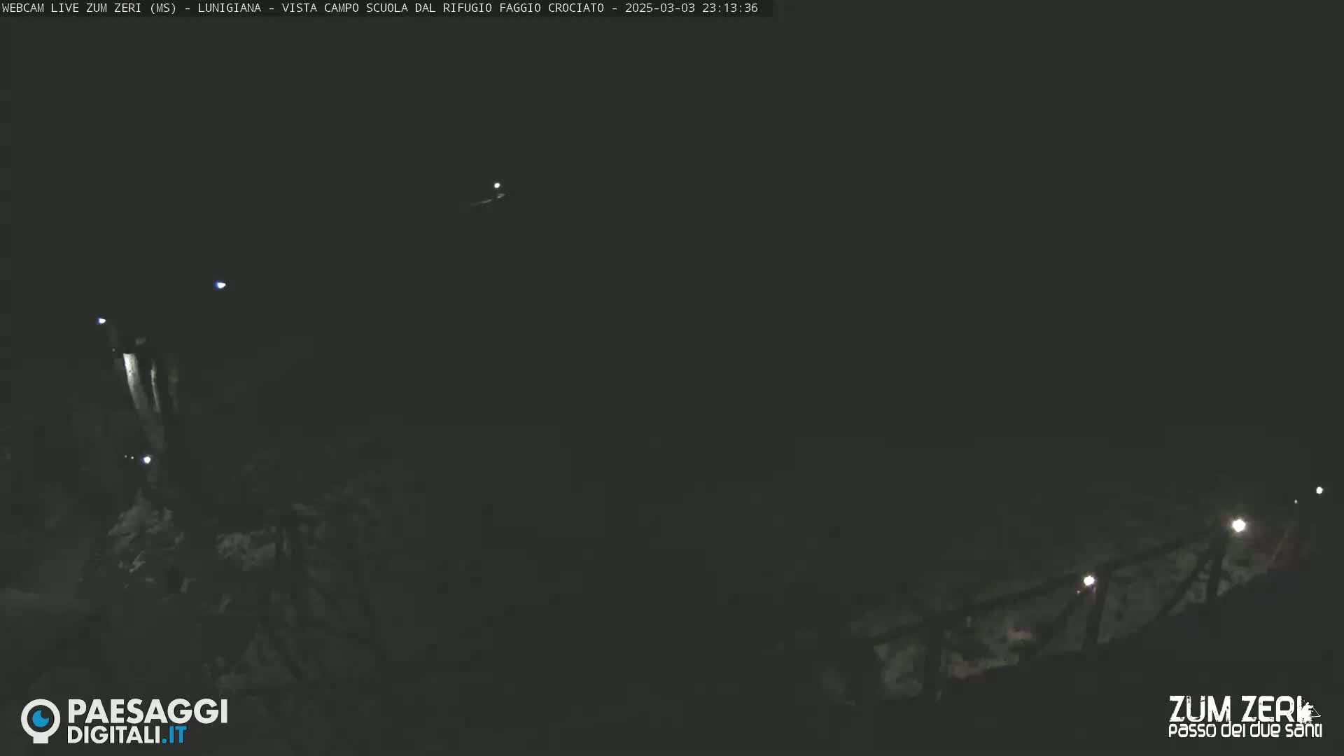 A nighttime, dark, outdoor scene shows a dimly lit path or road winding through a mountainous area.