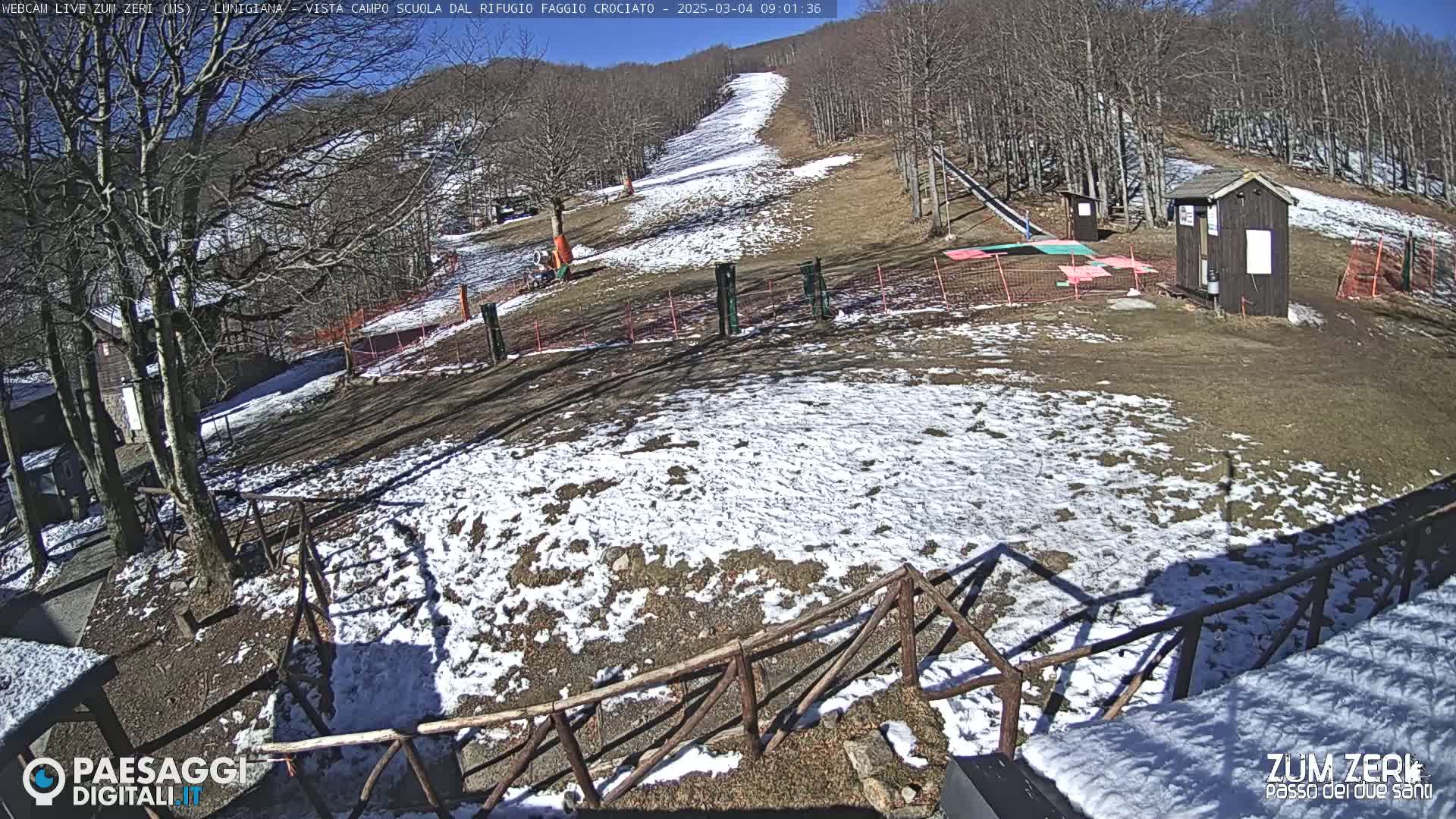 A sunny, snow-dusted ski slope with several small buildings and fencing is visible.