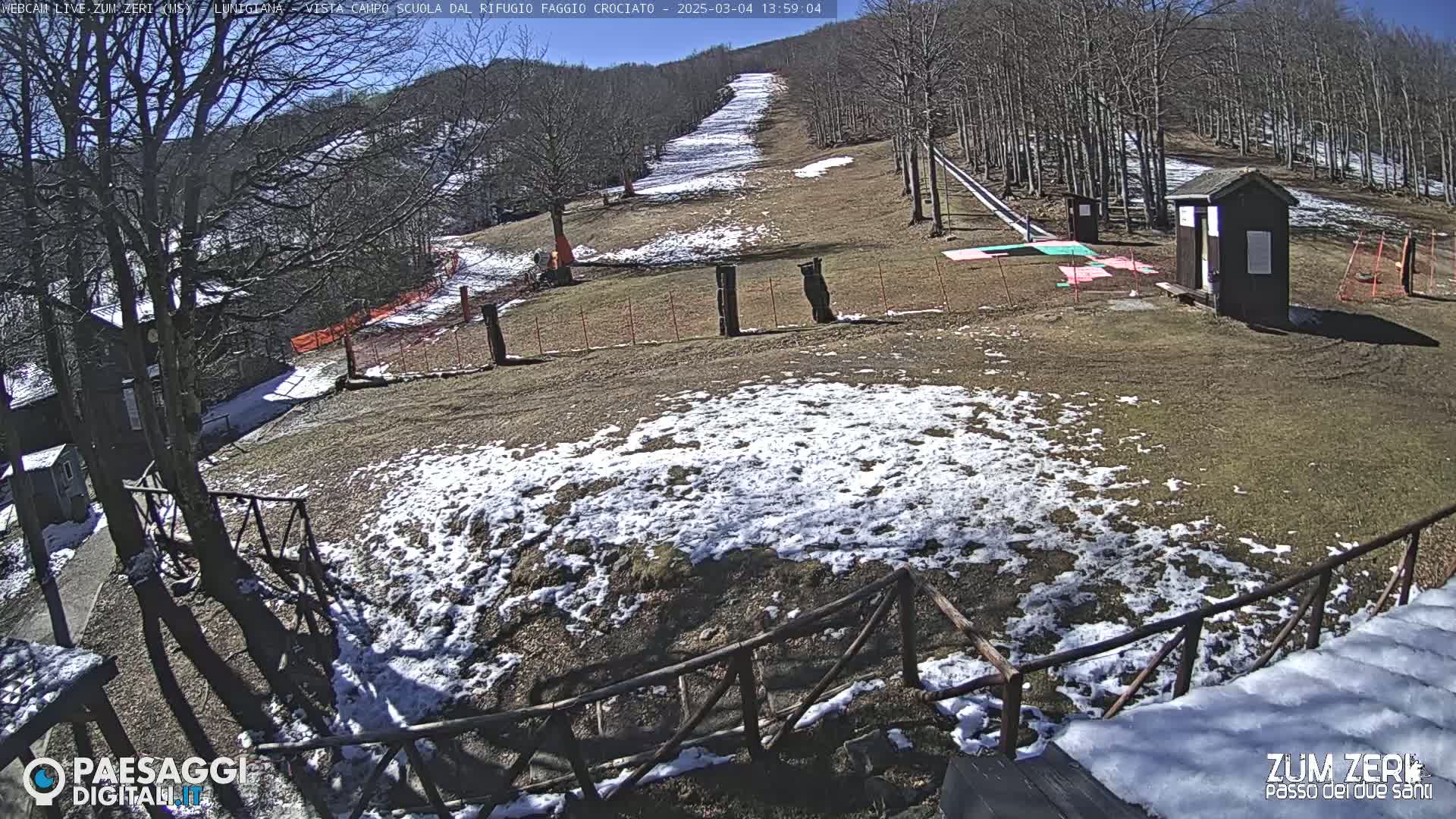 A sunny, partially snow-covered ski slope is visible, with a small building and several wooden posts near the bottom of the slope.