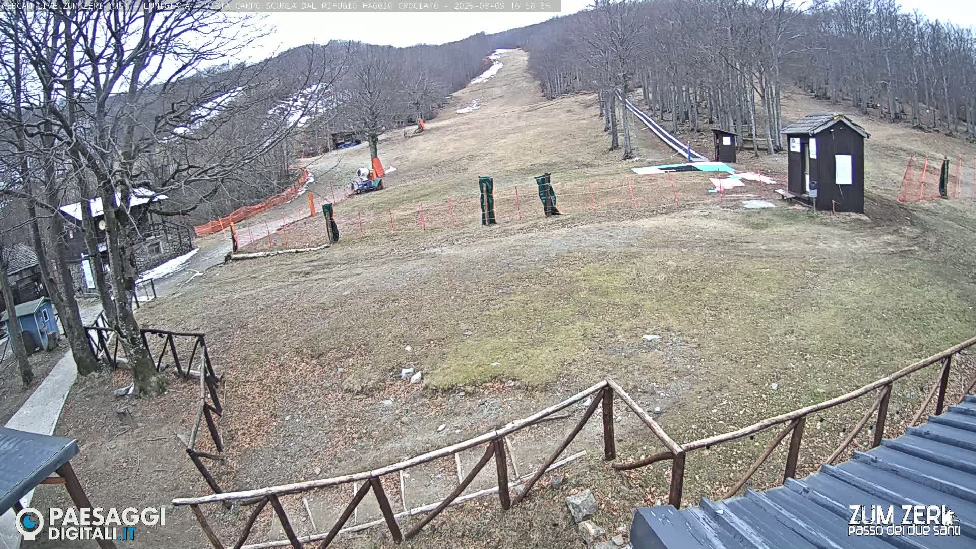 A mostly barren ski slope with a few buildings and a wooden fence is visible under an overcast sky.