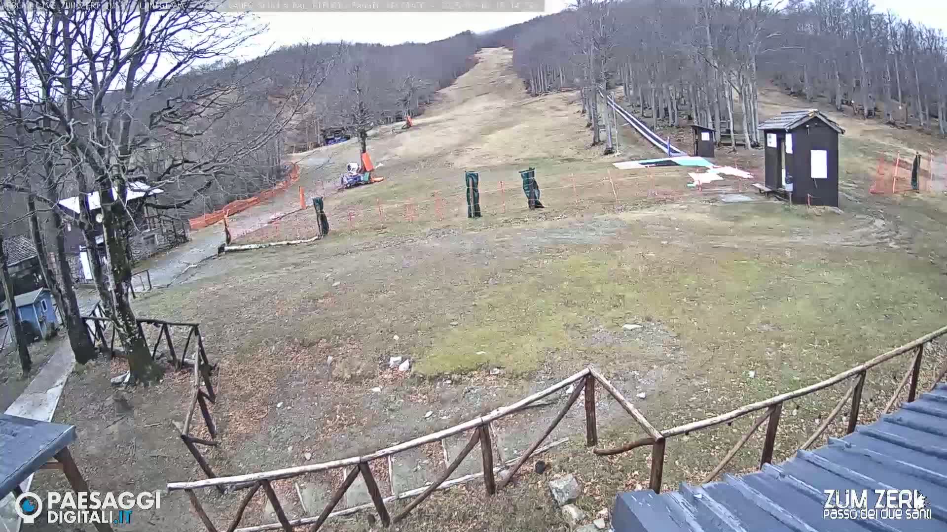 A mostly barren ski slope with a few small buildings and a wooden fence is visible under an overcast sky.