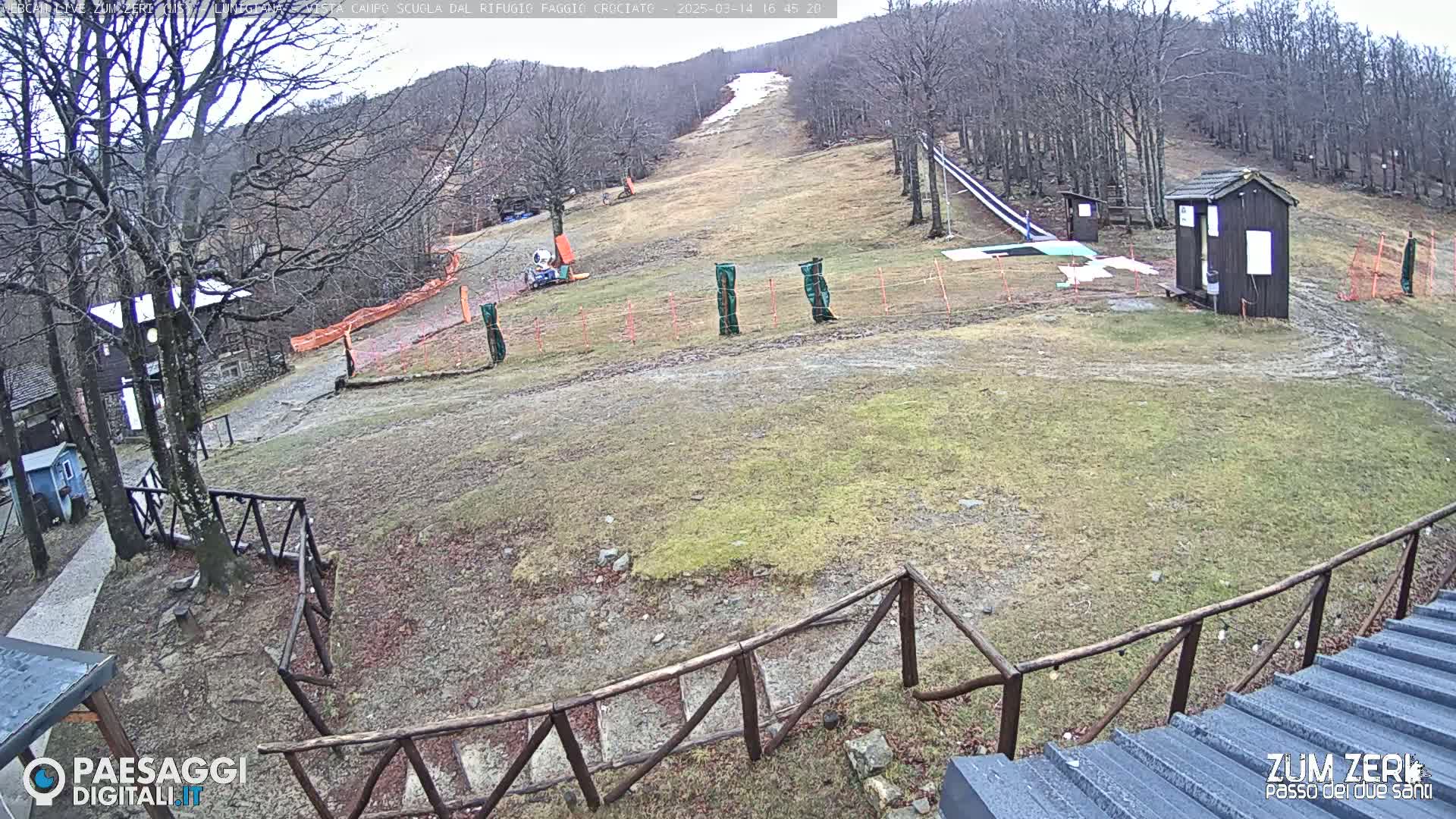 A mostly barren ski slope with a small building and a wooden fence is visible on an overcast day.