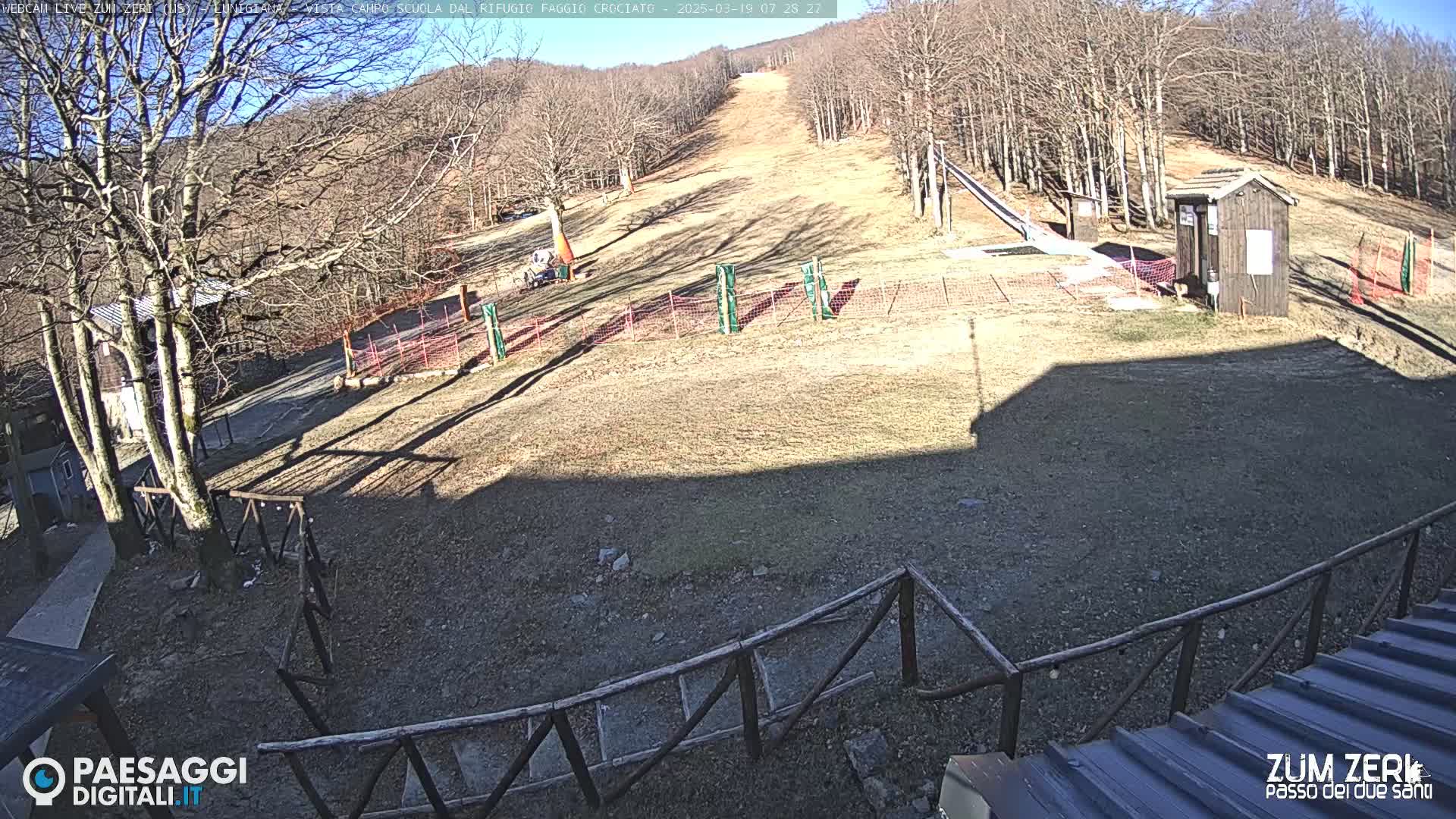 A sunny day at a ski slope shows a mostly barren hill with a small building, fencing, and a few trees in the background.