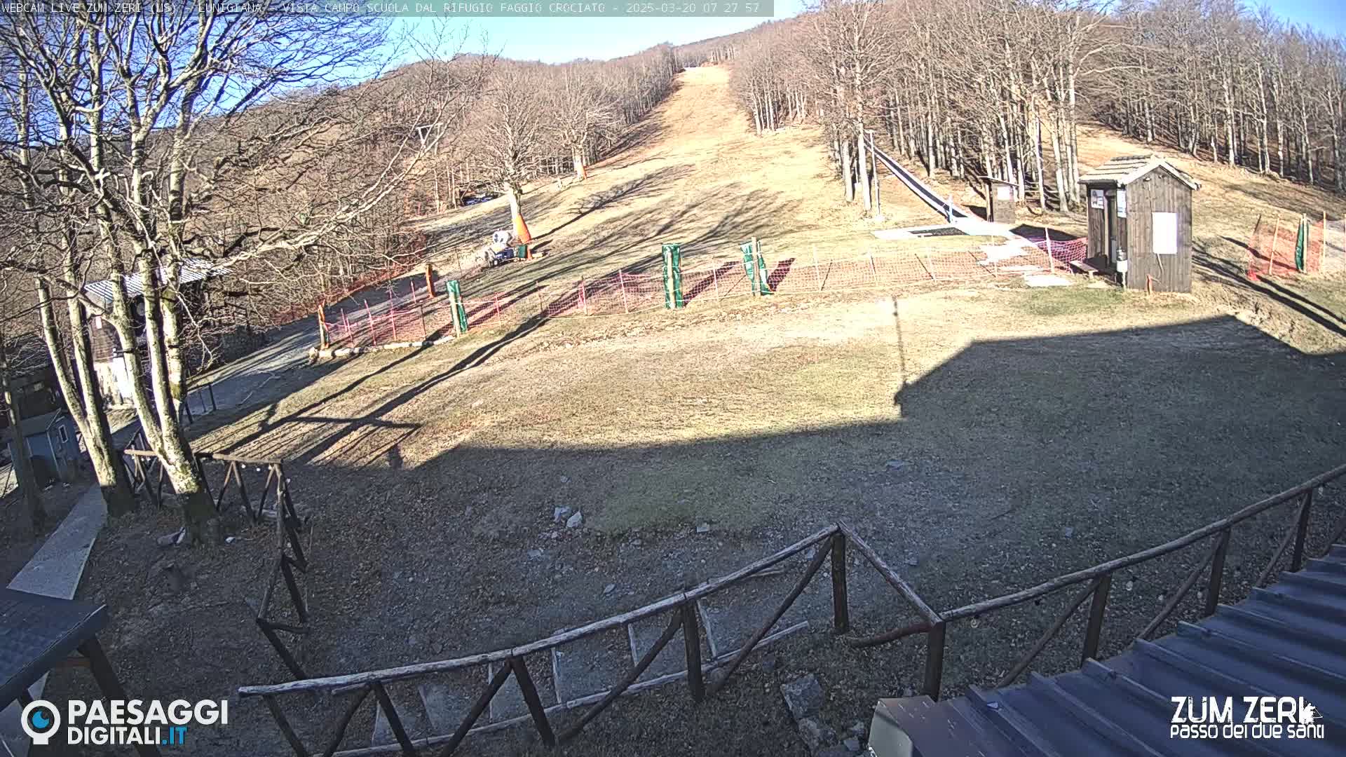 A sunny view of a ski slope with sparse snow, a small building, and several fences, surrounded by leafless trees.