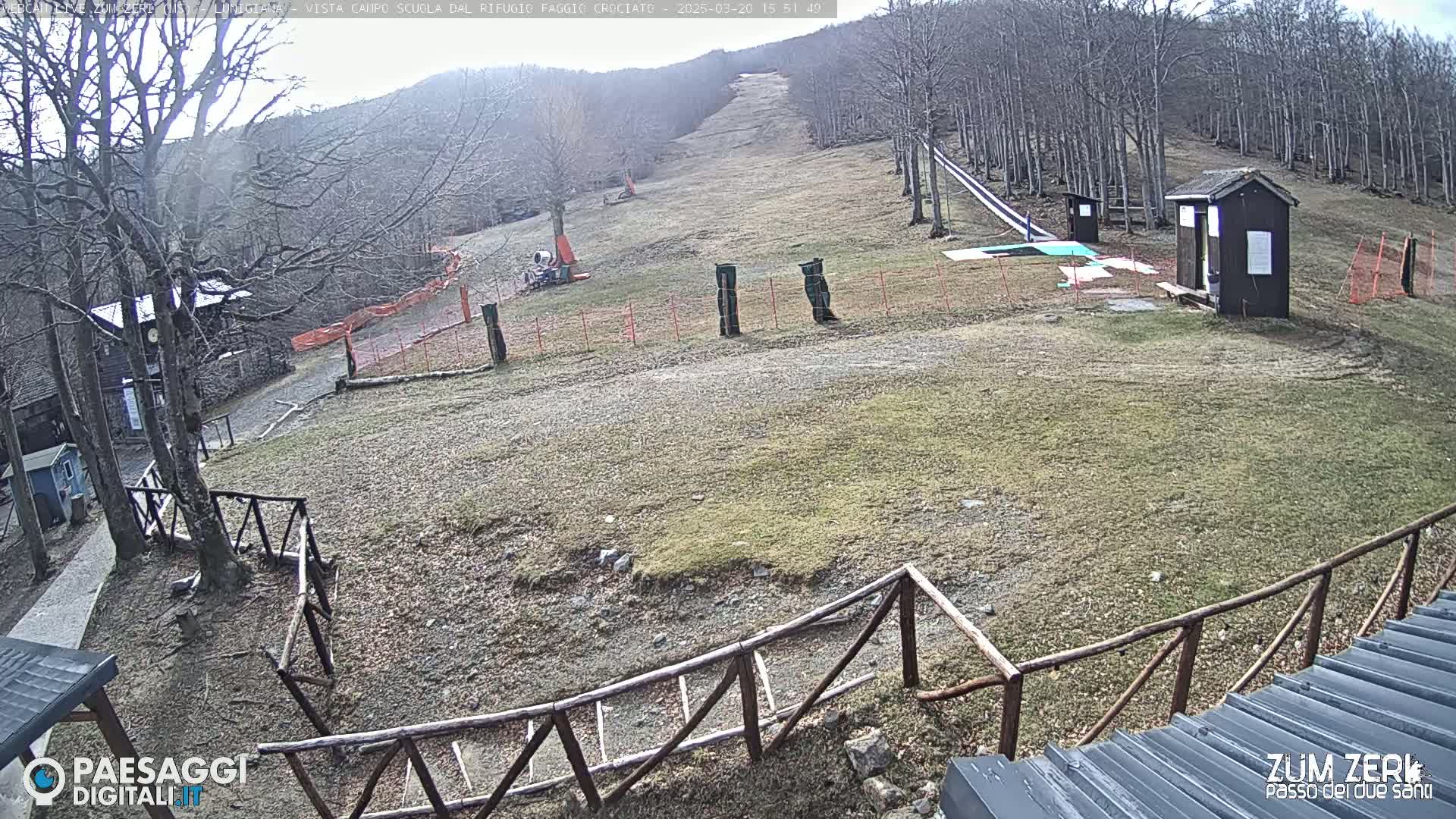 A mostly barren ski slope with small buildings and fences is visible, under a cloudy sky.