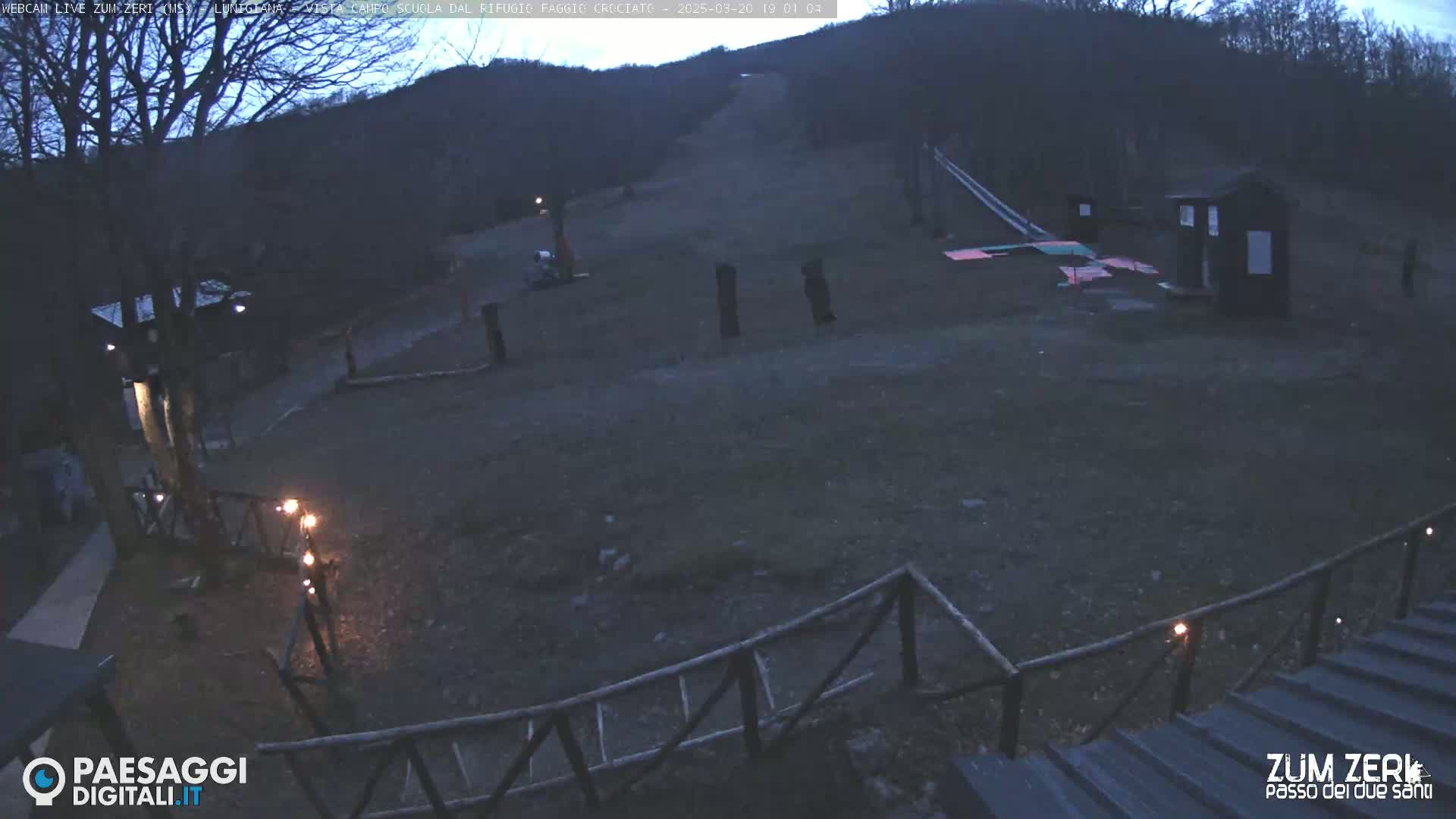 A dark, outdoor scene at dusk shows a sparsely populated area with a few small structures, a wooden fence, and a hill in the background.